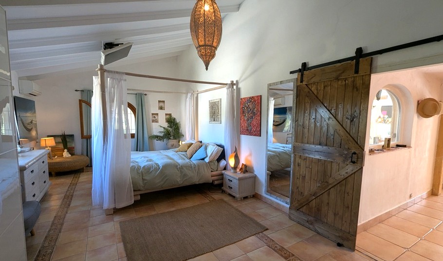Slaapkamer met hemelbed, wit balkenplafond en een rustieke houten schuifdeur. De terracotta vloer en Marokkaanse lamp versterken de mediterrane sfeer.