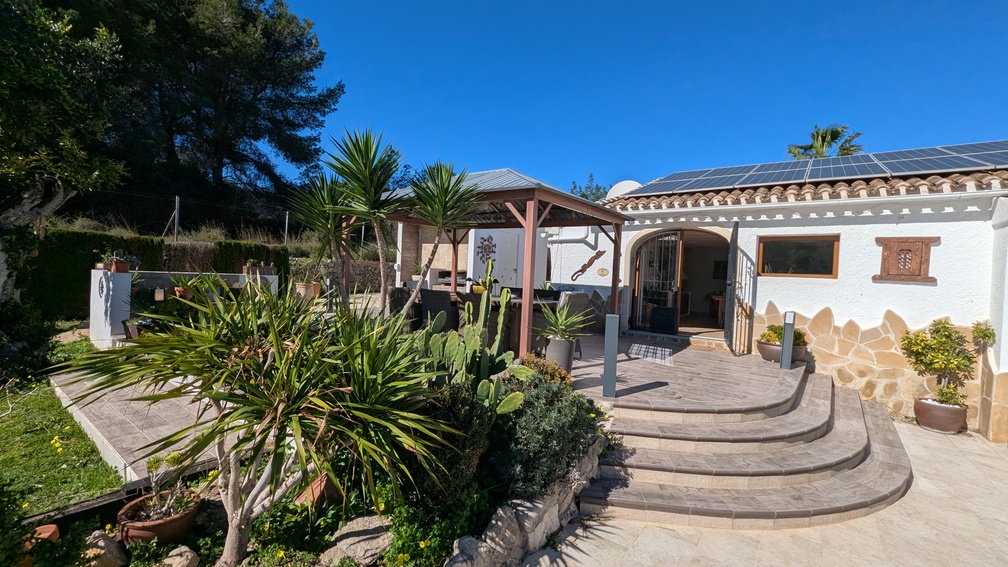 Terras met gebogen stenen trappen, een houten gazebo voor buitenmaaltijden en een witte gevel voorzien van zonnepanelen en mediterrane planten.