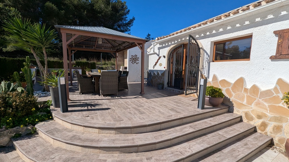 Verhoogd terras met tegels in steenlook en gebogen treden die leiden naar een schaduwrijke eethoek onder een houten pergola, grenzend aan een witte gevel met natuurstenen details.