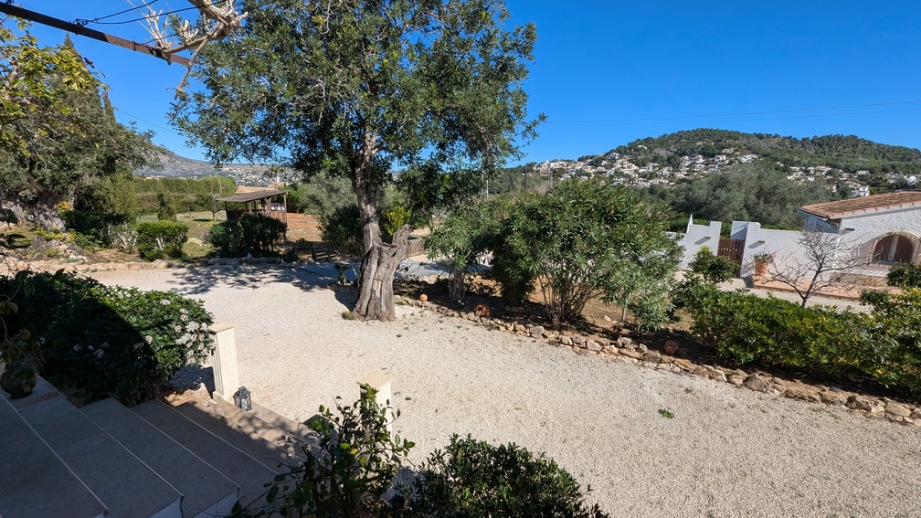 Verhoogd uitzicht op de grindoprit en mediterrane tuinen, met volwassen olijfbomen en uitzicht op de omliggende heuvels en woonwijken van Jávea.