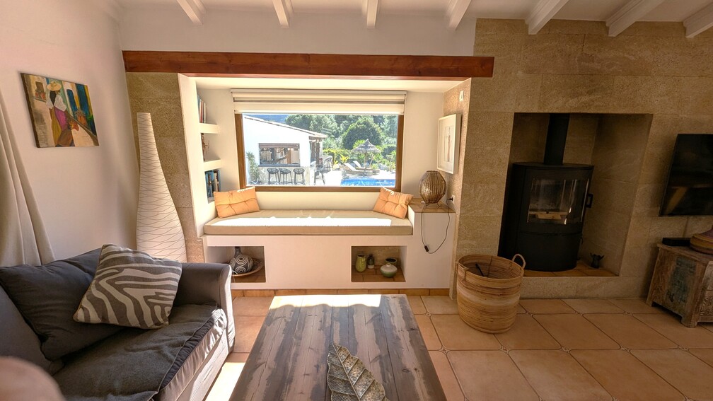 Woonkamer met ingebouwde vensterbank met uitzicht op het zwembad, moderne houtkachel in een wand van Tosca-natuursteen en balkenplafonds.