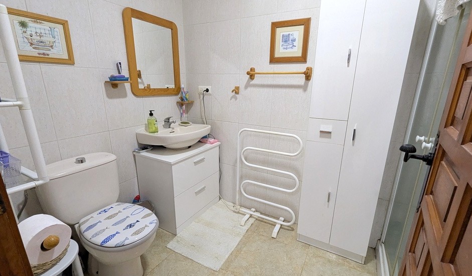 Functionele badkamer met wit wastafelmeubel, spiegel met houten lijst en opbergkasten. Voorzien van een handdoekradiator en betegelde wanden.