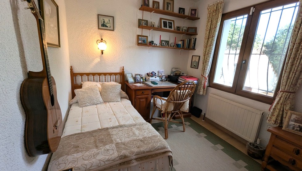 Slaapkamer met houten eenpersoonsbed, klassiek bureau, rieten stoel en wandplanken. Voorzien van een radiator en raam met beveiligingsroosters.
