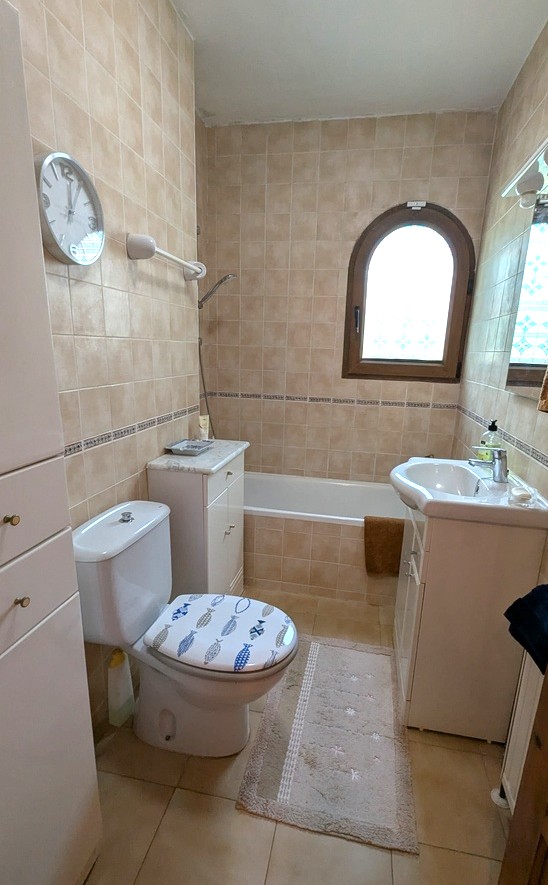 Traditionele badkamer met beige betegeling, een ingebouwd ligbad en een houten boogvenster. Voorzien van een wit wastafelmeubel en extra opbergruimte.