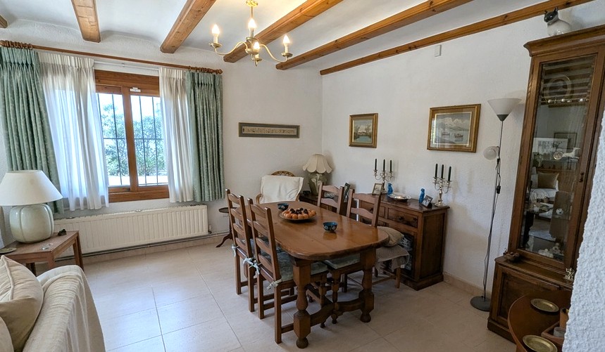 Traditionele eethoek met houten plafondbalken, een massief houten tafel en een klassiek dressoir. De ruimte heeft een radiator en een raam met beveiligingsroosters.
