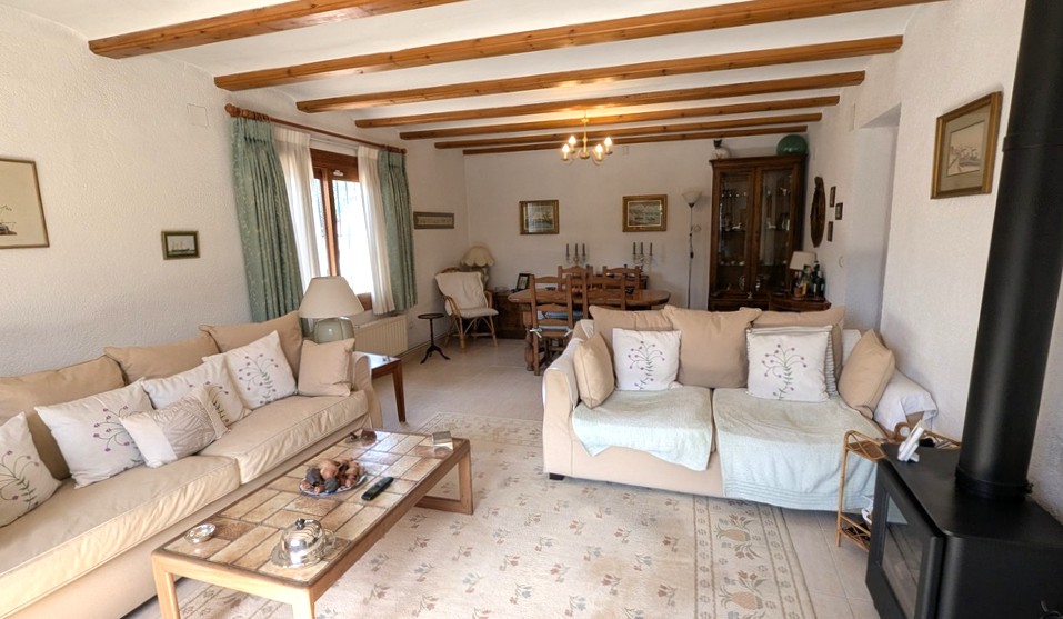 Traditionele woon- en eetkamer met zichtbare houten plafondbalken, een zwarte houtkachel en klassiek meubilair in neutrale tinten.