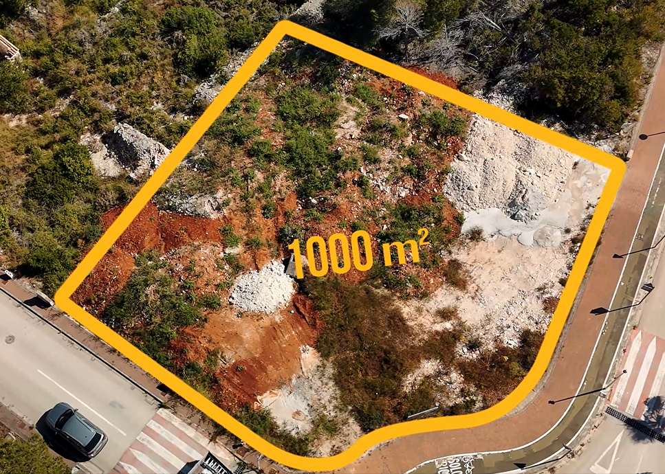 Luchtfoto van het hoekperceel van 1.000 m² in Jávea, met de kenmerkende rode aarde en ligging aan een volledig verharde weg met trottoirs en straatverlichting.