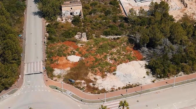 Luchtfoto van het hoekperceel van 1.000 m² in Villas del Vent, met rode aarde, omringende pijnbomen en een volledig geasfalteerde infrastructuur.