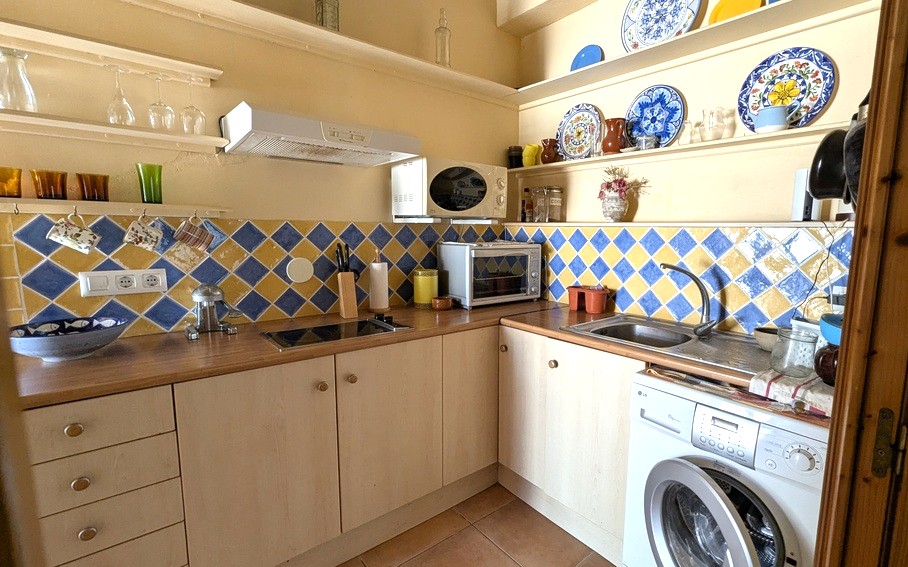 Compacte keuken met blauw-gele ruitvormige tegels, houten werkbladen en open planken met traditioneel keramiek. Inclusief wasmachine en diverse inbouwapparatuur.