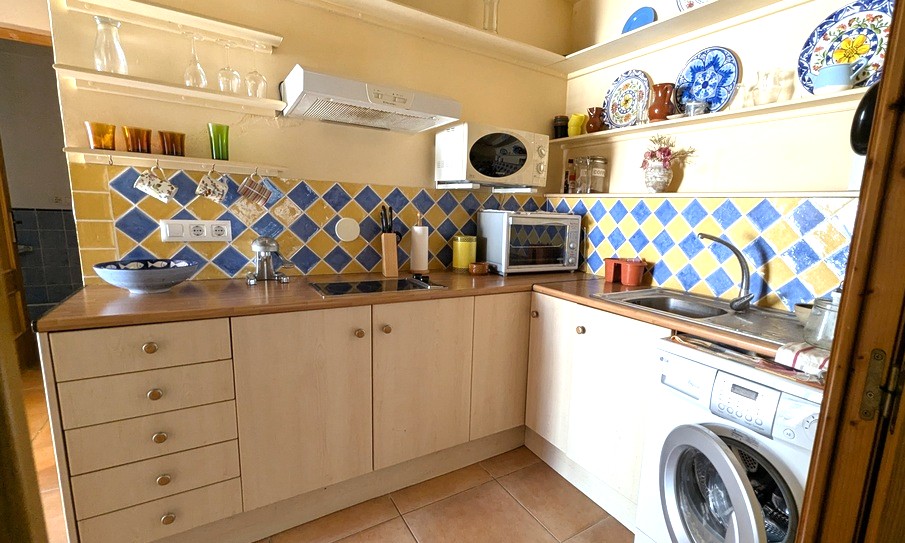 Compacte L-vormige keuken met een blauw-gele ruitvormige tegelwand, houten werkbladen, open schappen en een ingebouwde wasmachine.