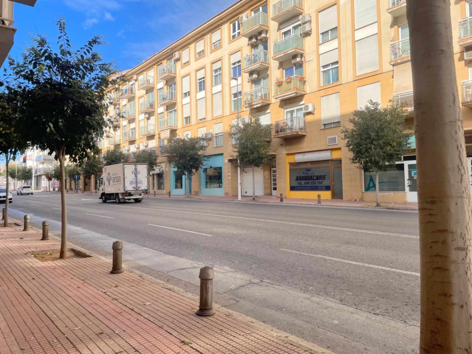 Buitenaanzicht van de Avenida Palmela in Jávea, met de gevel van het gebouw, commerciële ruimtes op de begane grond en brede trottoirs met bomen.