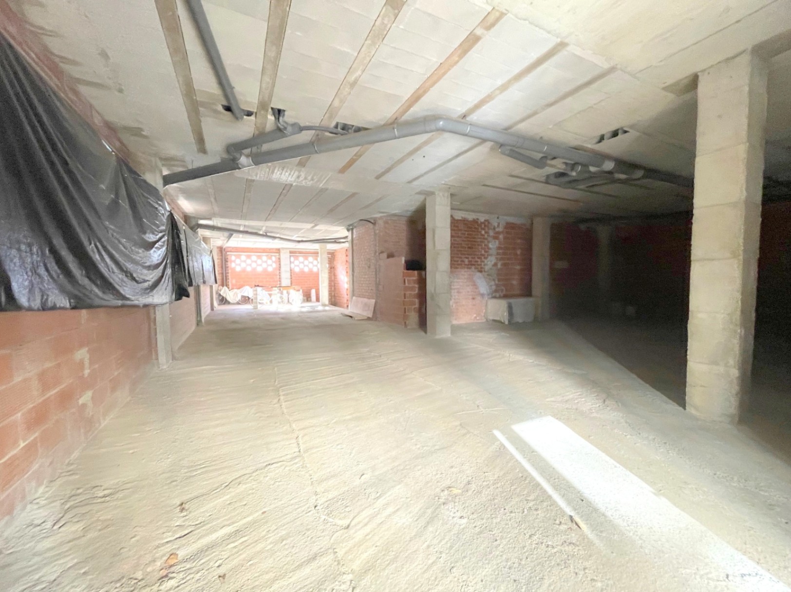 Open commerciële ruimte in ruwbouwstaat met betonnen kolommen, zichtbare plafondleidingen en bakstenen muren die een flexibele indeling bieden.