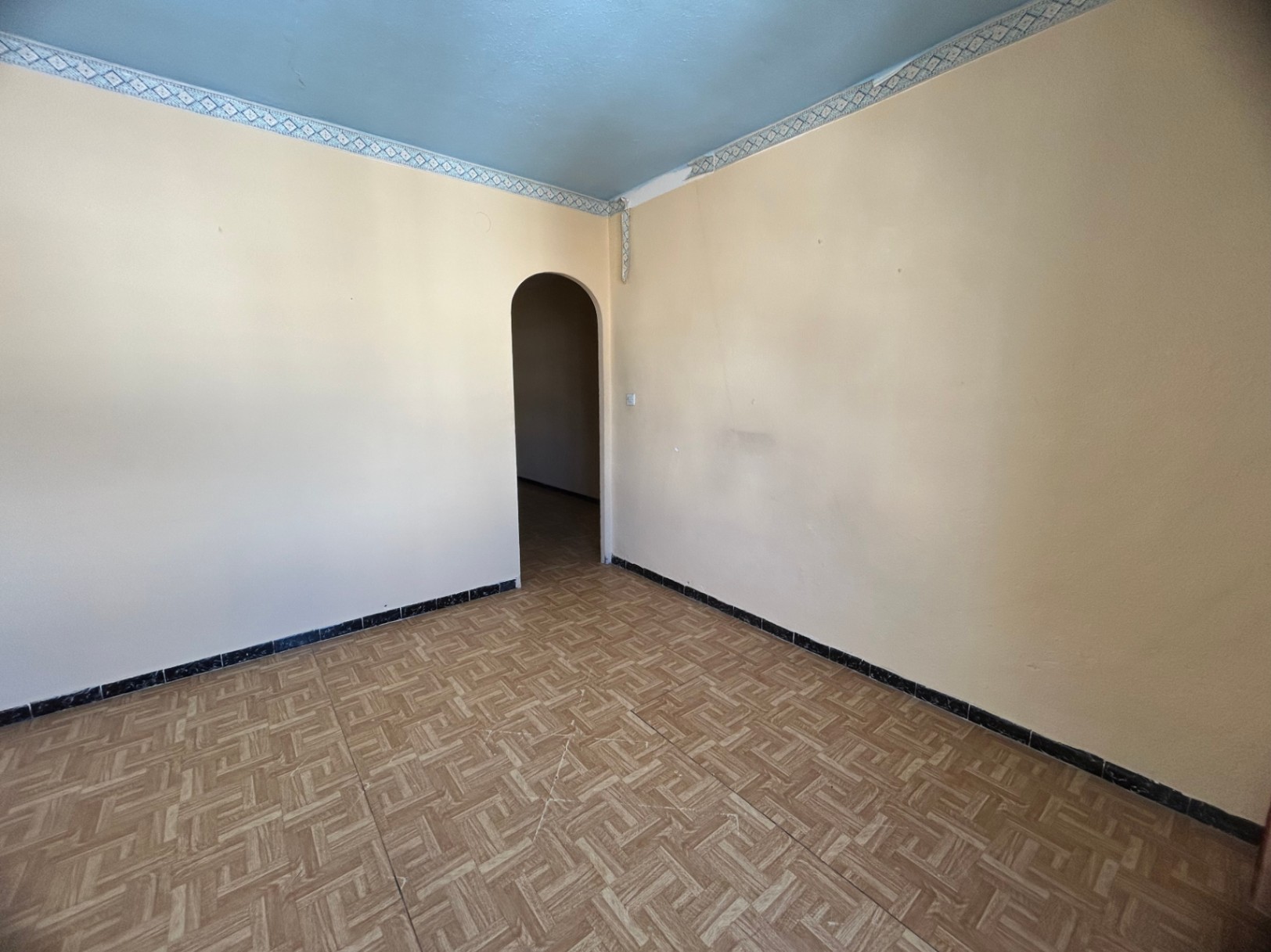 Binnenruimte met gedessineerde keramische tegelvloer, crème muren en een lichtblauw plafond met sierrand. Een ronde toog biedt toegang tot de volgende kamer.