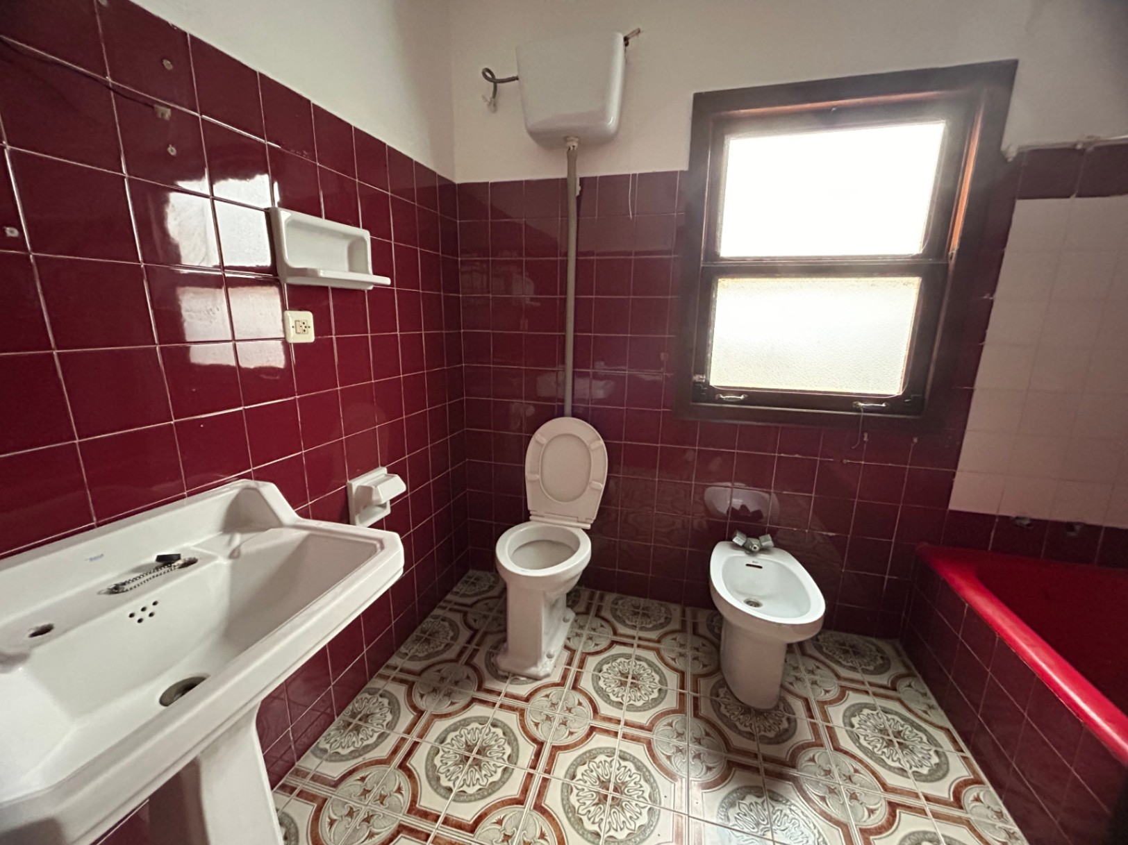 Originele badkamer met bordeauxrode wandtegels, gedecoreerde hydraulische vloer en klassiek sanitair. Voorzien van een houten raam voor natuurlijk licht en ventilatie.