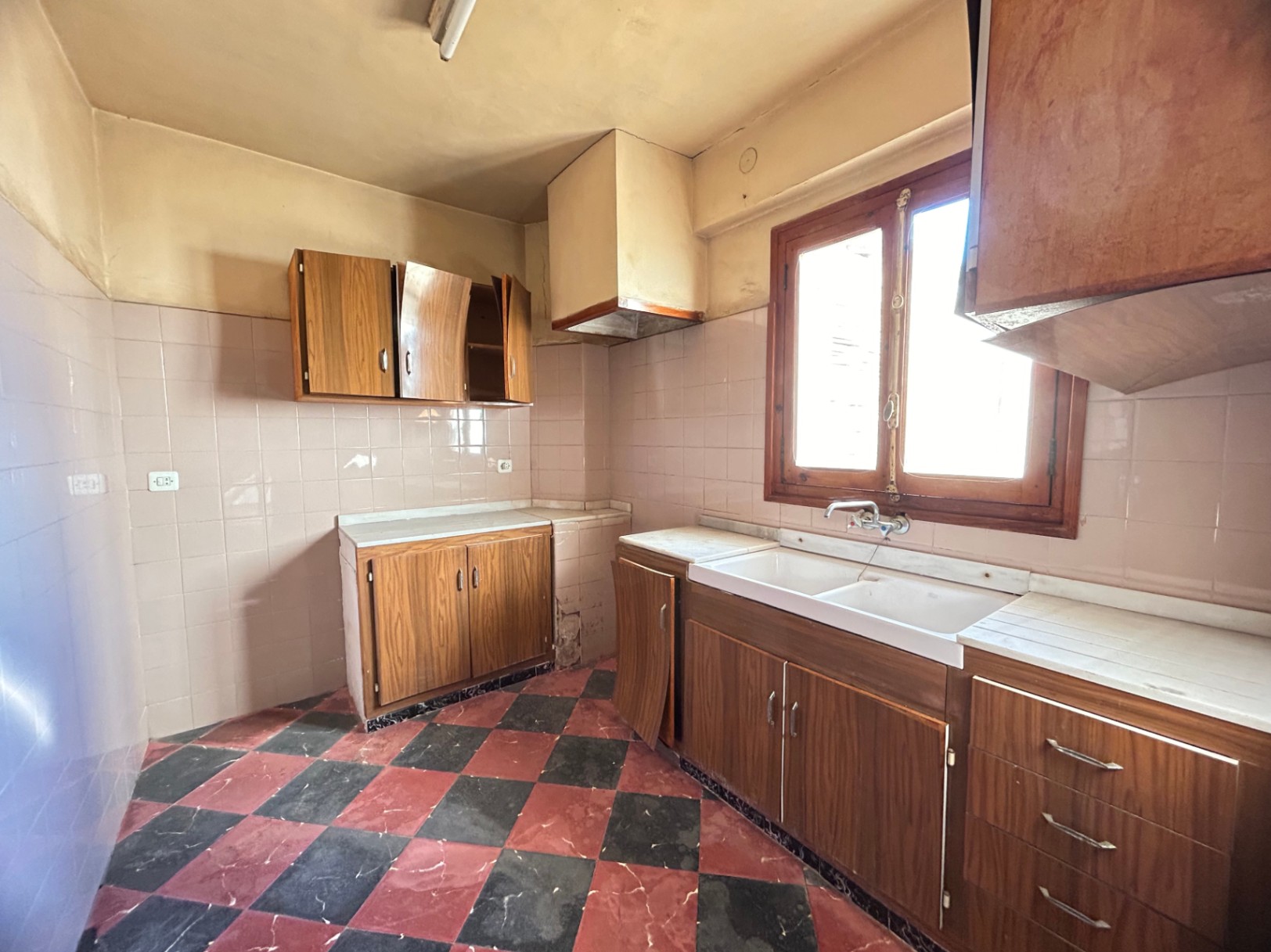 Originele keuken met rood-zwarte dambordvloer, houten kasten en wit marmeren werkbladen. Voorzien van een dubbele spoelbak en een houten raamkozijn.