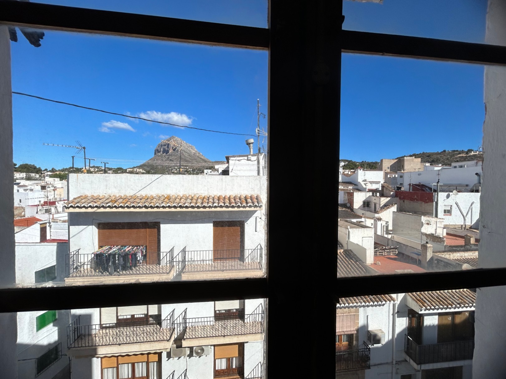 Stedelijk uitzicht van binnenuit door een raamkozijn, uitkijkend over de daken van de oude stad van Jávea met de top van de Montgó-berg in de verte.