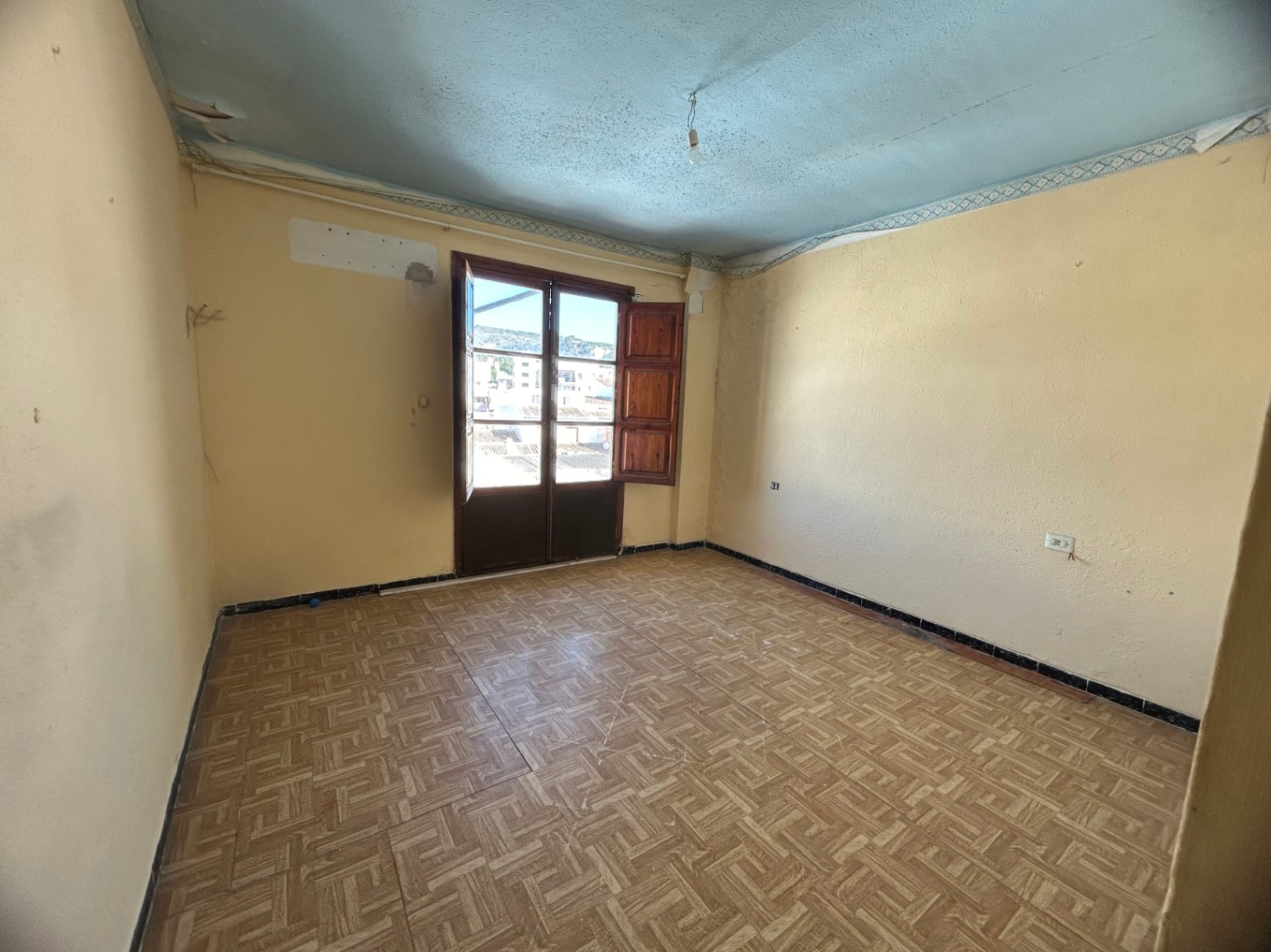 Te renoveren kamer met keramische tegelvloer in patroon, crèmekleurige wanden en houten openslaande deuren naar een balkon met uitzicht op de stad.