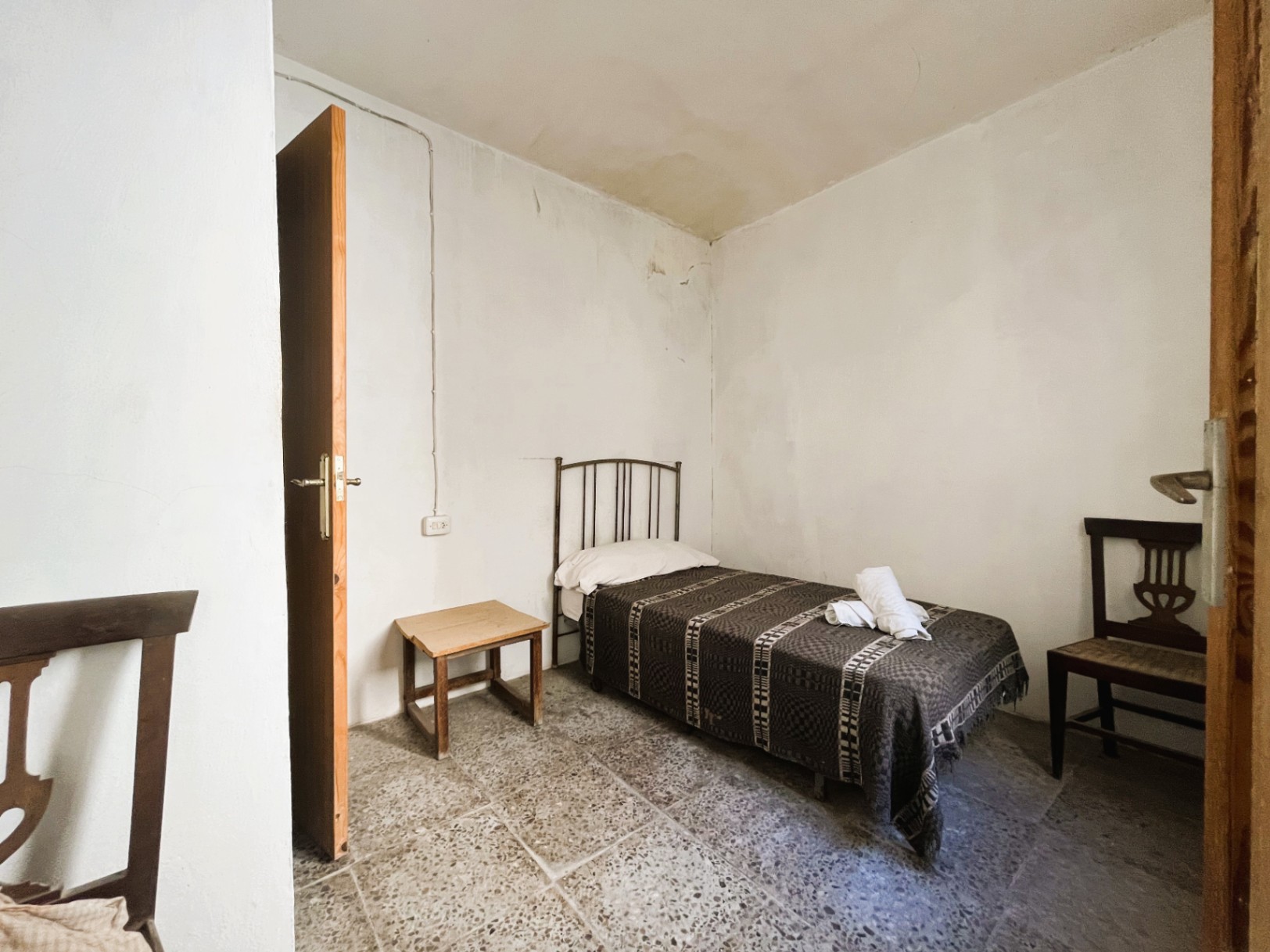 Eenvoudige slaapkamer met originele terrazzovloer, eenpersoonsbed met metalen hoofdbord en traditionele houten meubels in een kamer met witte muren.