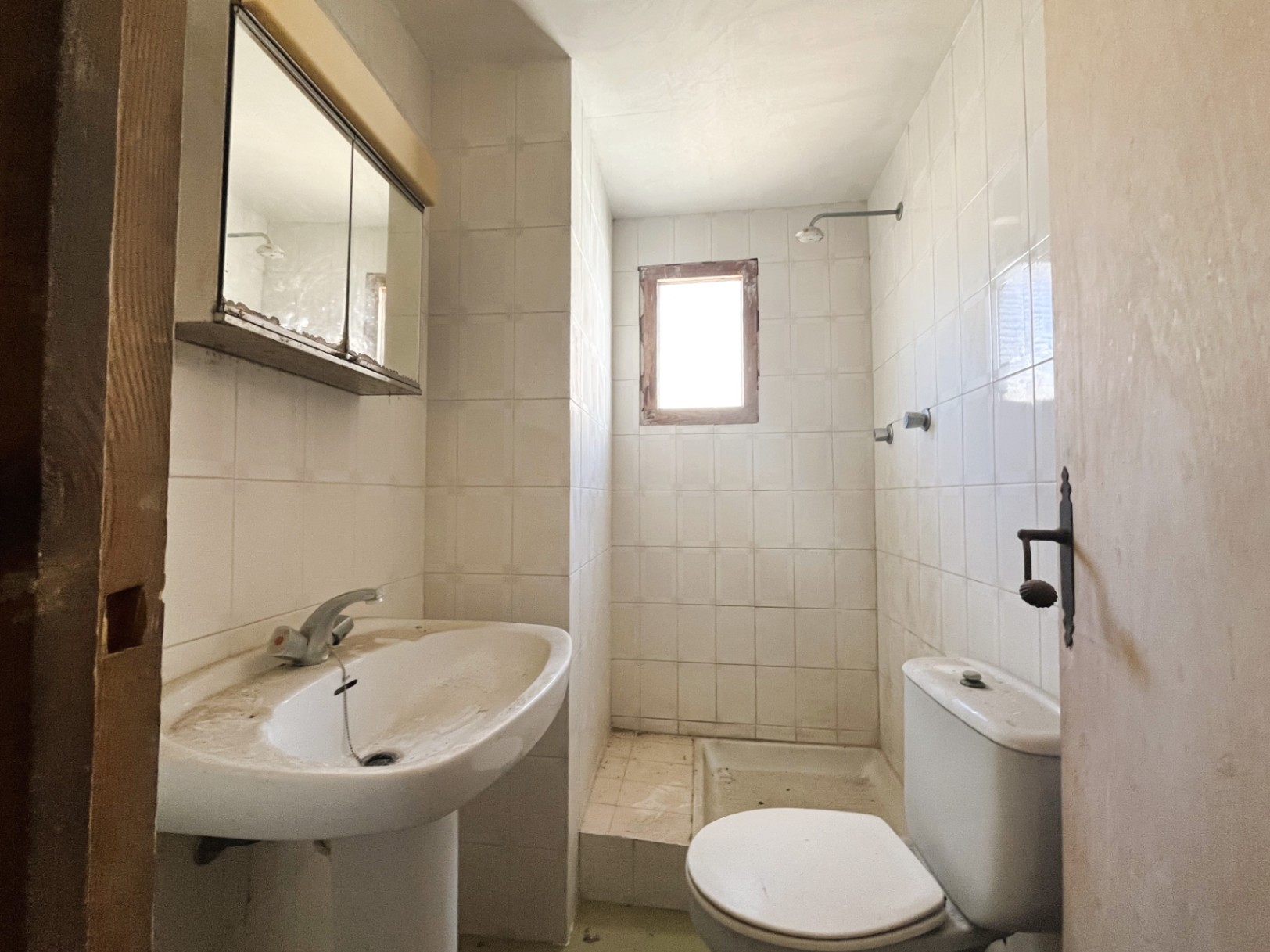 Functionele badkamer met witte tegels, een wastafel op zuil en doucheruimte met een klein raam. Een ruimte met veel potentieel voor renovatie.