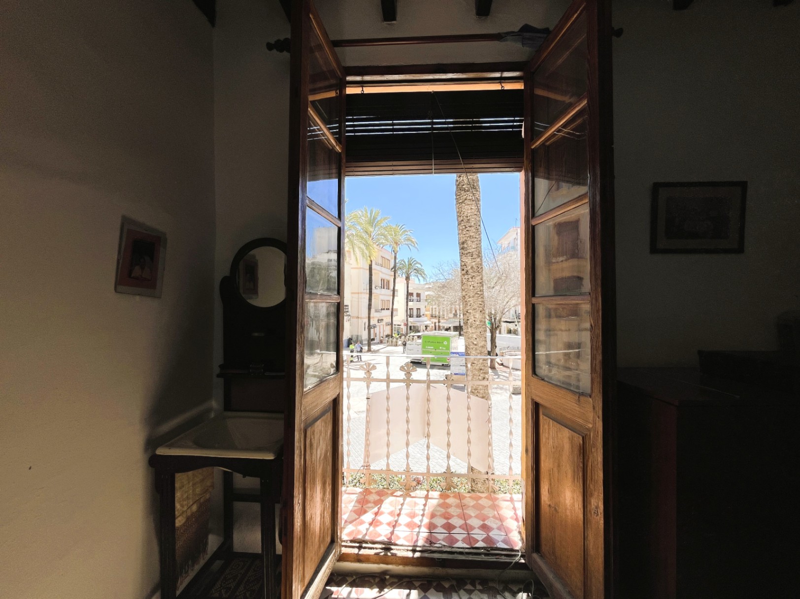 Binnenzicht door houten deuren naar een balkon met rood-witte ruitjesvloer en uitzicht op een zonnig plein met palmbomen in het historische centrum.