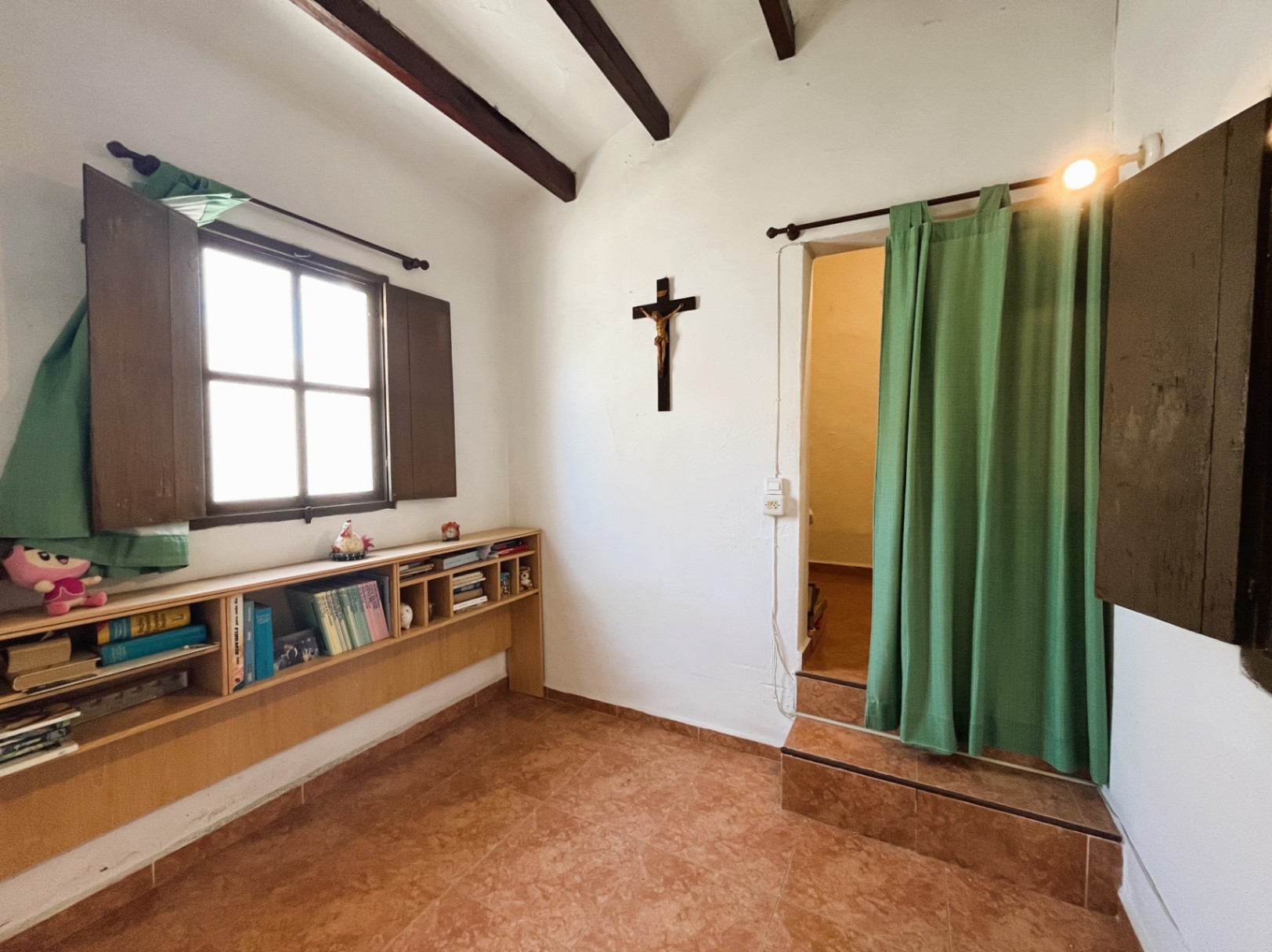 Interieur met donkere houten balken, terracotta vloer en een lage boekenkast. Een verhoogde doorgang met treden en een groen gordijn biedt toegang tot de volgende ruimte.