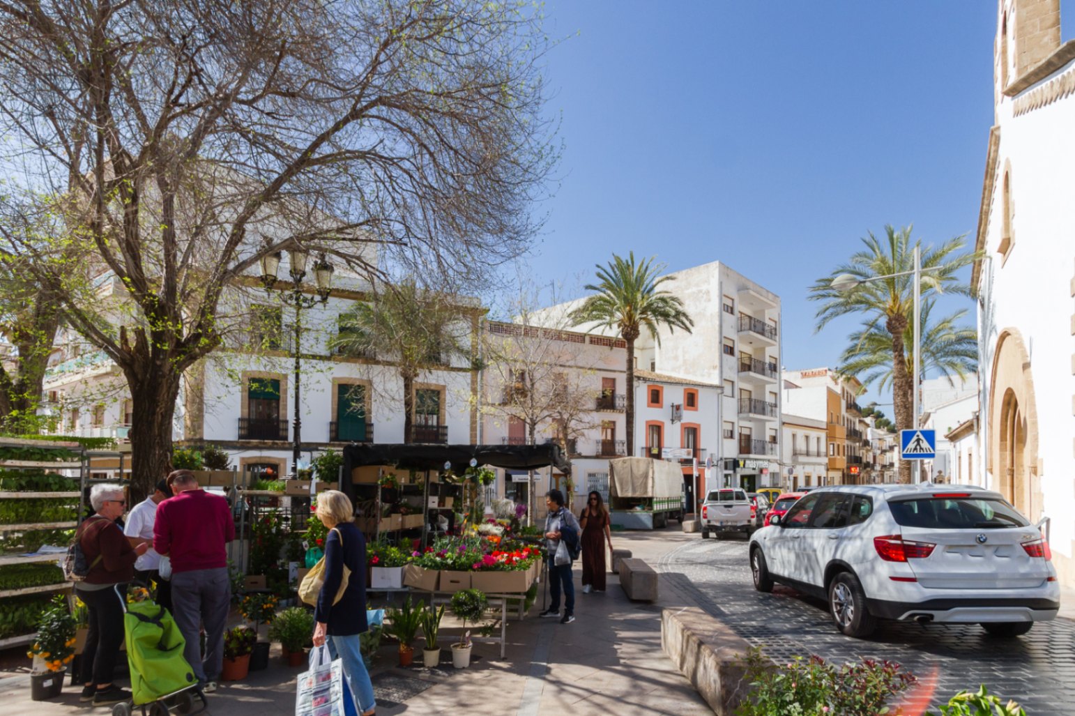 Plein in het historische centrum van Jávea met een bloemenkraam, omgeven door traditionele witte architectuur, kasseien en palmbomen in het volle daglicht.