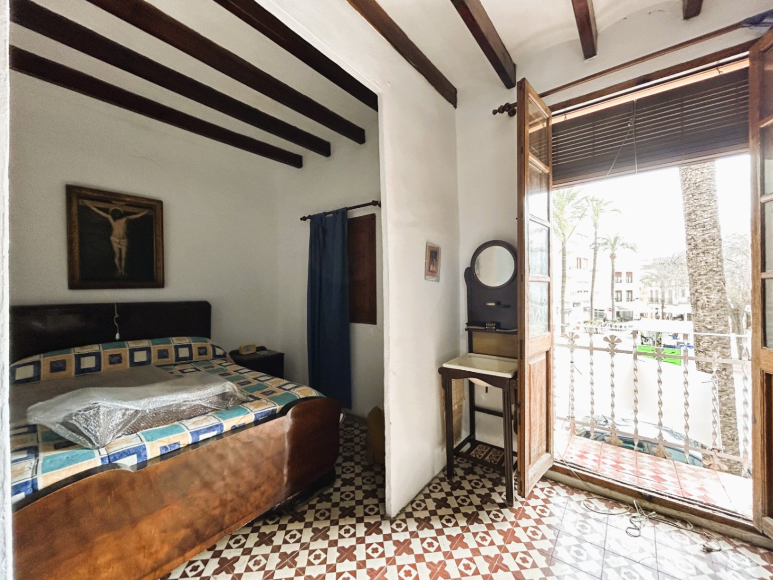 Traditionele slaapkamer met houten balkenplafond, authentieke patroontegels en een balkon met uitzicht op het dorpsplein en palmbomen.