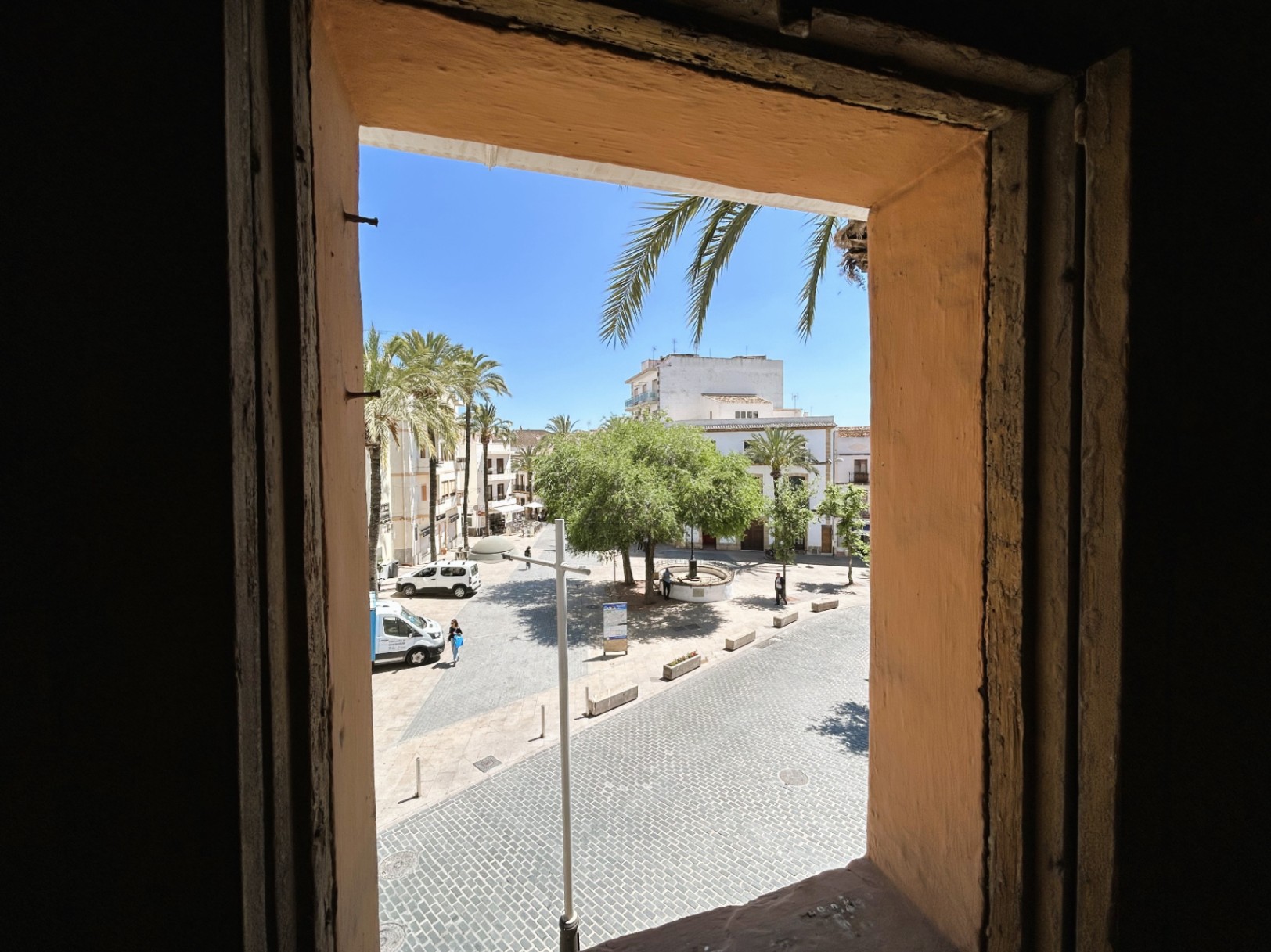 Uitzicht vanuit het interieur op een geplaveid plein in het oude centrum, met de nadruk op de traditionele dikke muren en de zonnige ligging met palmbomen.