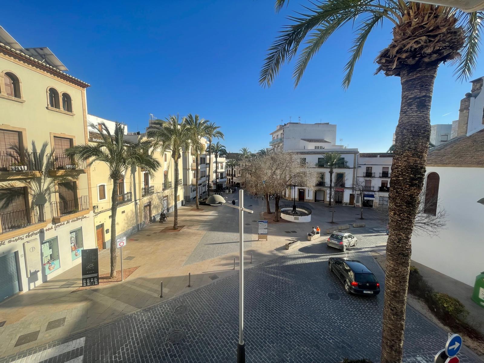 Verhoogd uitzicht vanaf de woning op een geplaveid plein in het oude centrum van Jávea, omgeven door traditionele mediterrane gebouwen en palmbomen.