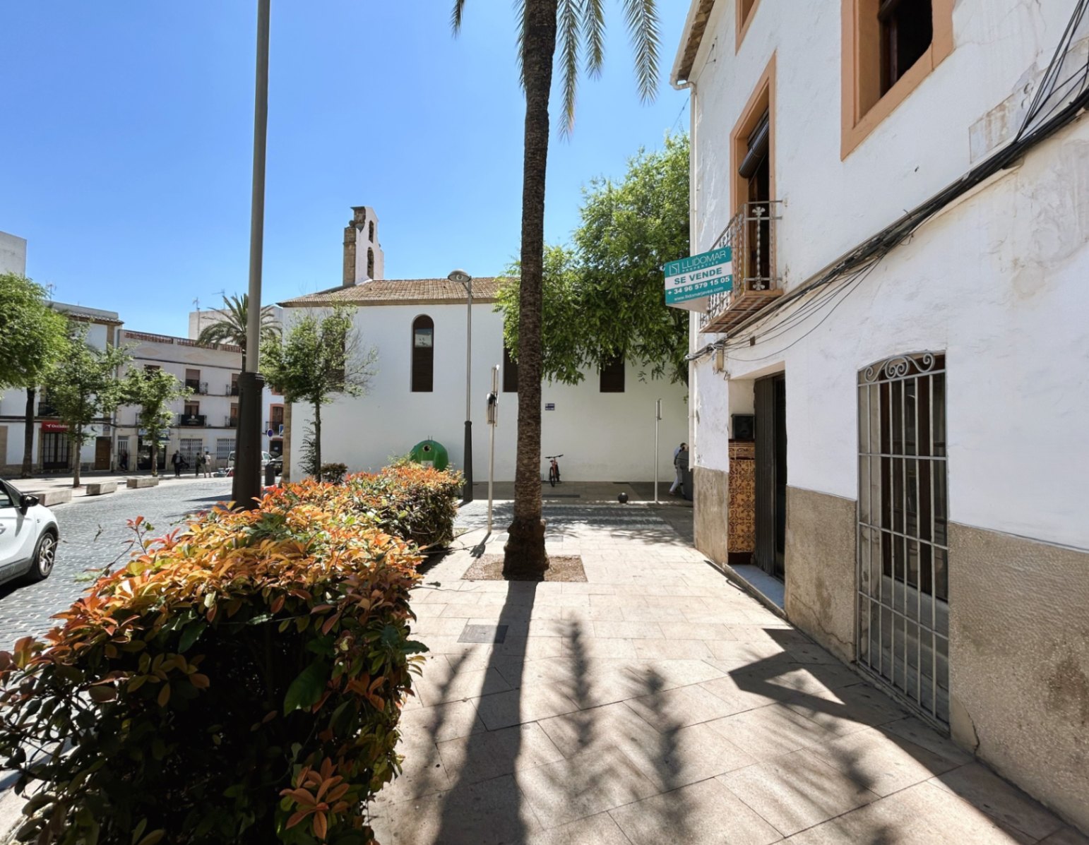 Witte gevel met stenen plint in de oude binnenstad van Jávea, gelegen aan een voetgangersplein met palmbomen en uitzicht op een historische klokkentoren.