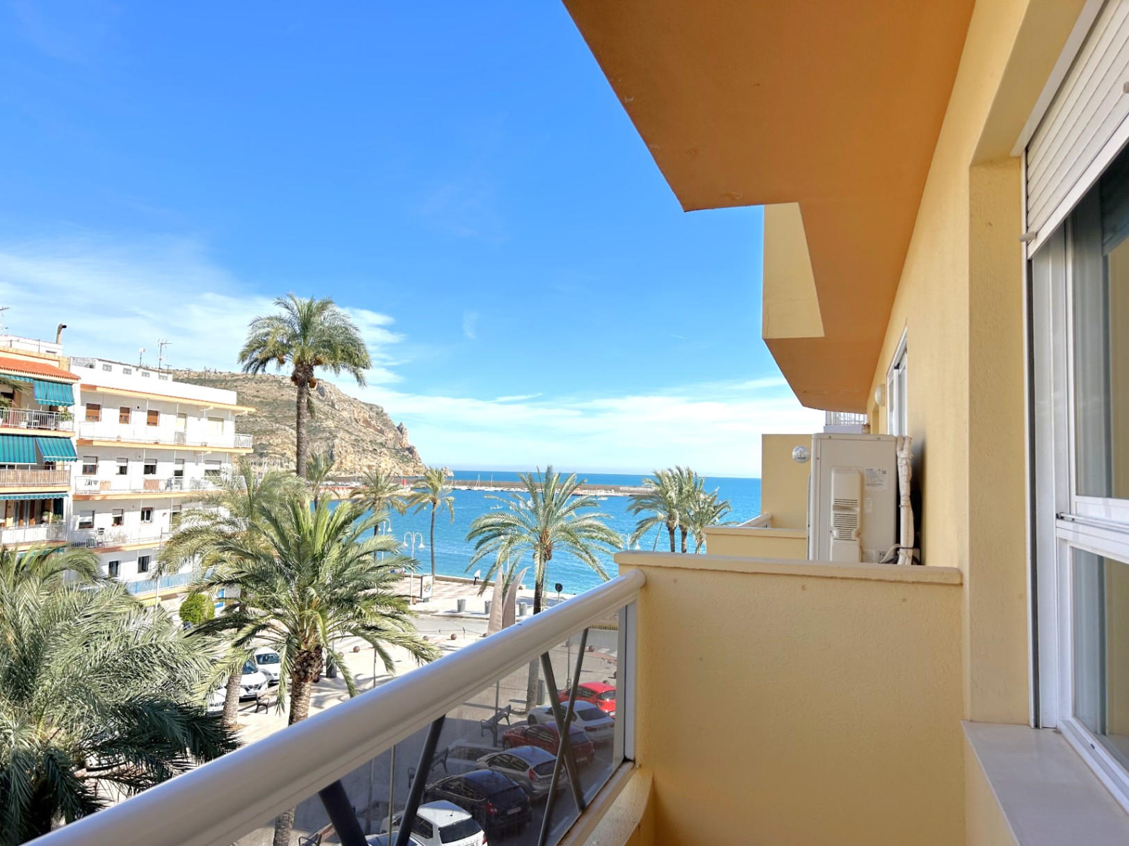 Balkon met direct uitzicht op de zee en de haven van Jávea, met palmbomen, een metalen reling en witte aluminium buitenkozijnen.