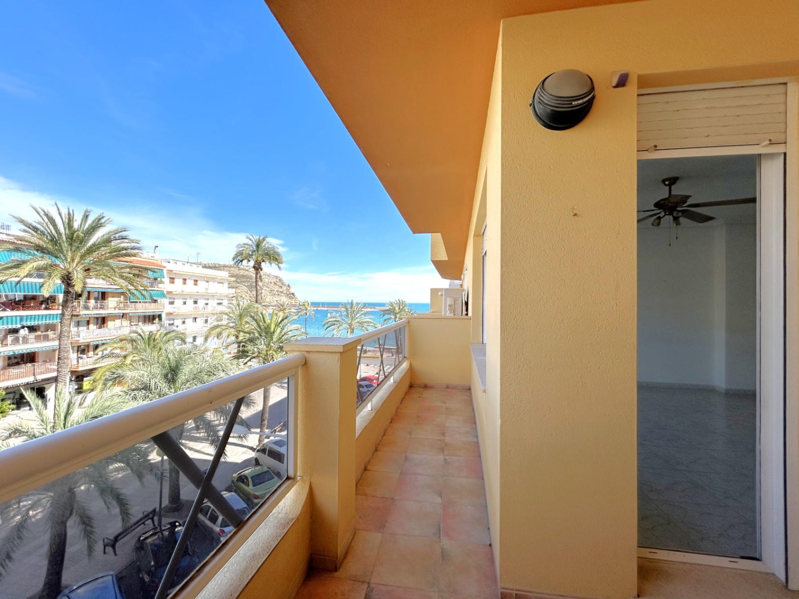 Langwerpig balkon met keramische tegels en zijdelings uitzicht op de Middellandse Zee en de haven van Jávea, omgeven door palmbomen.