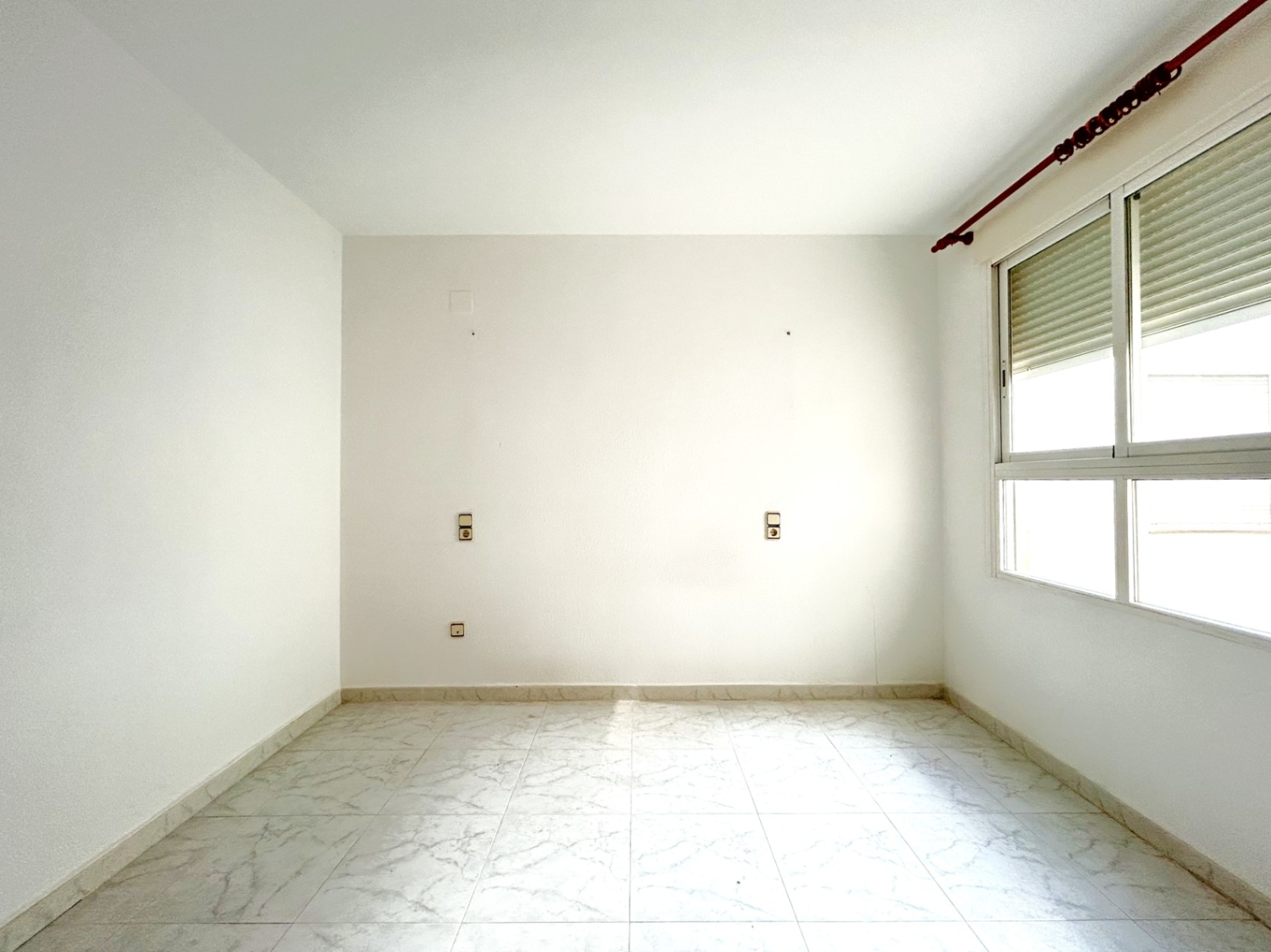 Lege slaapkamer met marmerlook tegelvloer, witte muren en een raam voorzien van een rolluik. Symmetrische stopcontacten zijn geplaatst voor de indeling van het bed.