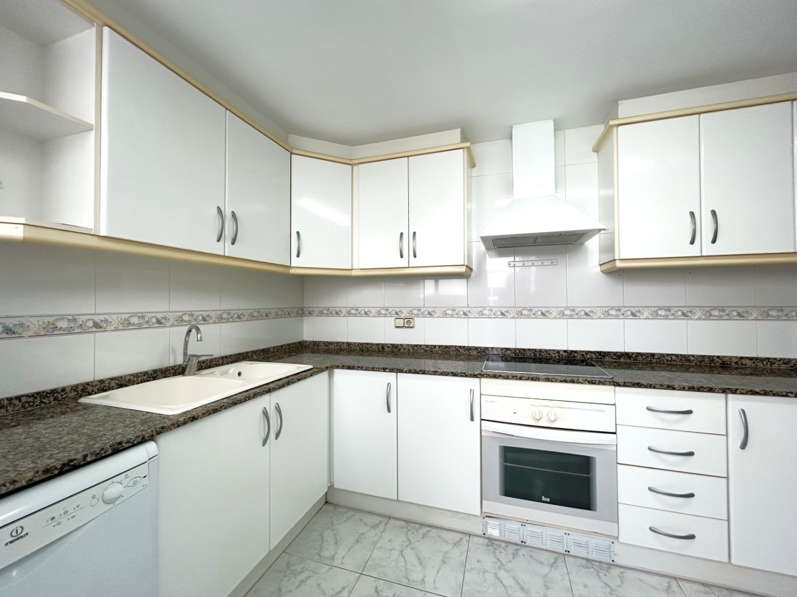 Onafhankelijke keuken met witte kasten, granieten werkblad, keramische kookplaat en oven. De wanden zijn betegeld en voorzien van een decoratieve sierstrip.