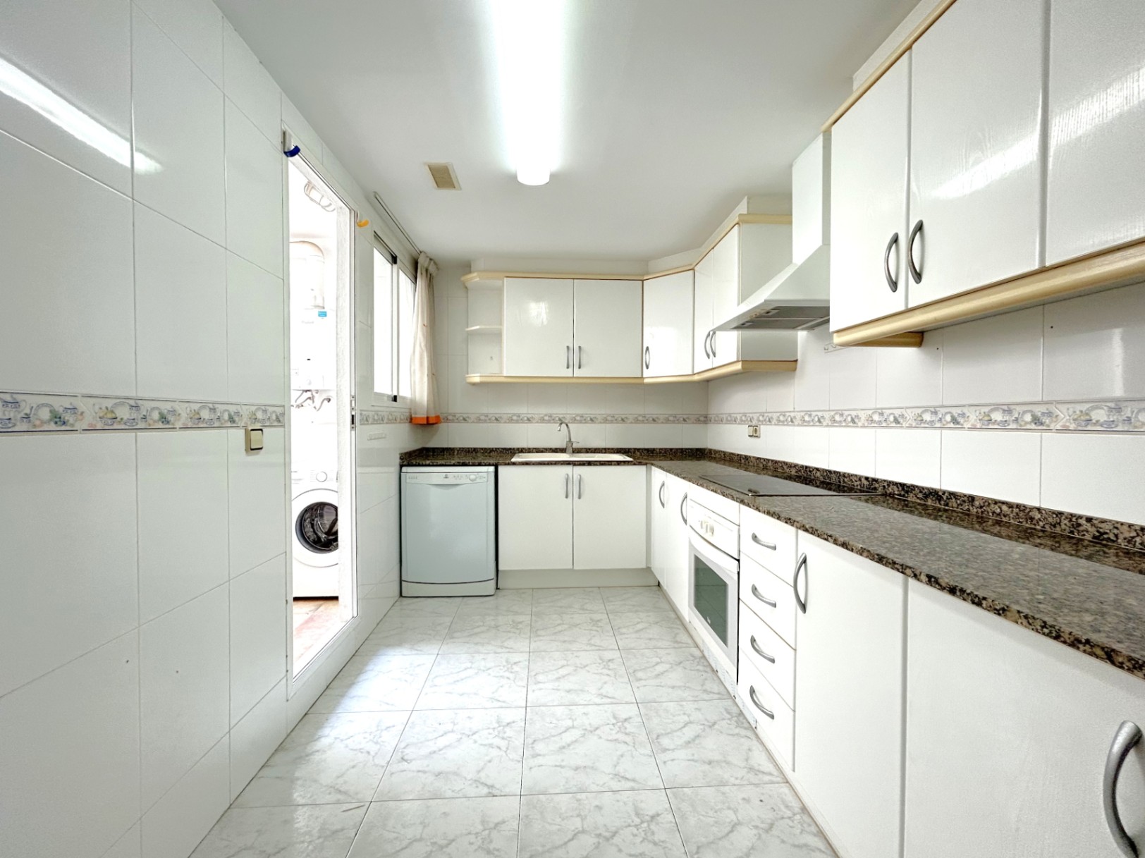 Onafhankelijke L-vormige keuken met witte kasten, donker granieten werkblad en toegang tot een aangrenzende wasruimte.