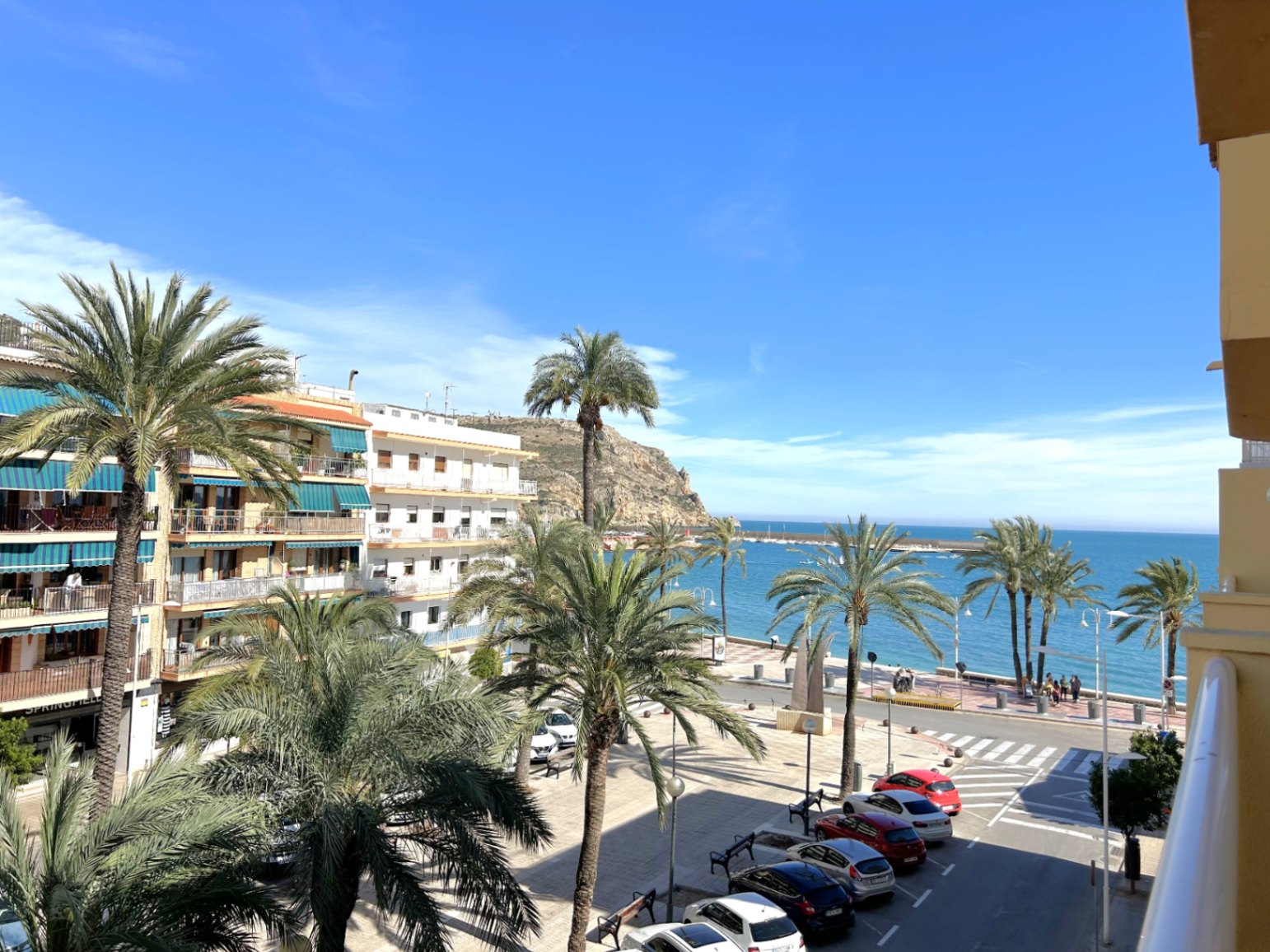 Uitzicht vanaf het balkon op de boulevard van de haven van Jávea, met palmbomen, de Middellandse Zee en de kustrotsen onder een heldere hemel.