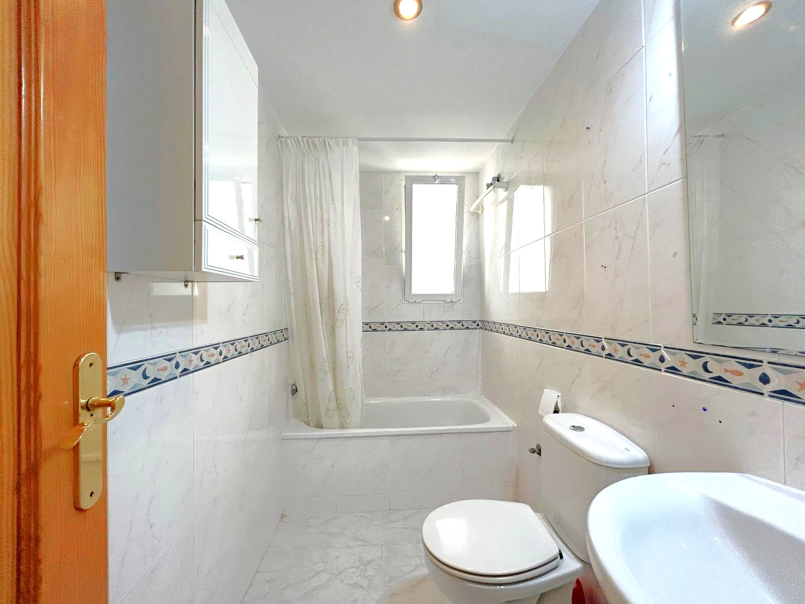 Volledige badkamer met ligbad, tegels met marmerlook en een decoratieve rand. Voorzien van een raam voor natuurlijk licht en functionele witte kasten.