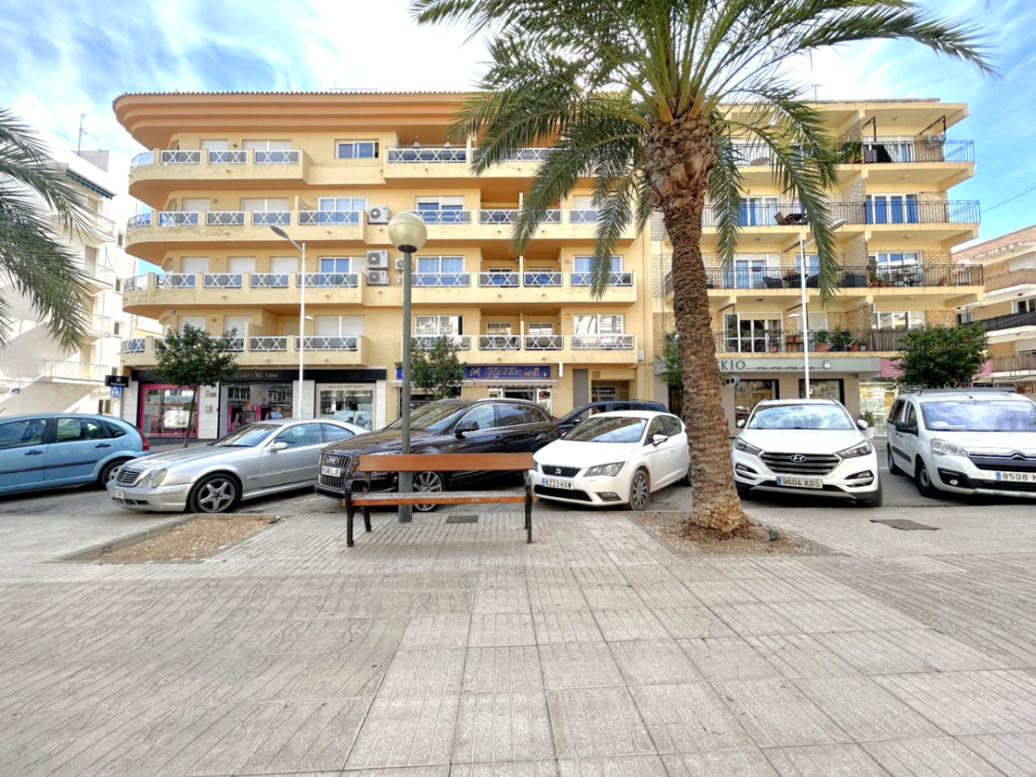 Vooraanzicht van het appartementencomplex in de haven van Jávea, met een okergele gevel en balkons met witte hekwerken aan een plein met palmbomen.