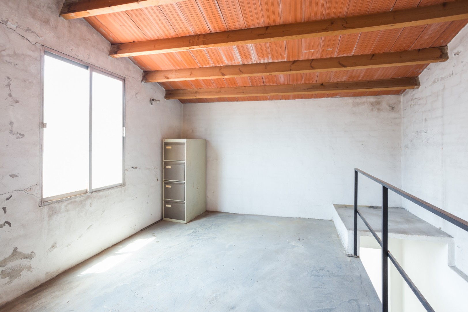 Bovenverdieping met zichtbare houten balken, een plafond van kleitegels, betonnen vloer en een groot raam voor veel natuurlijk licht.
