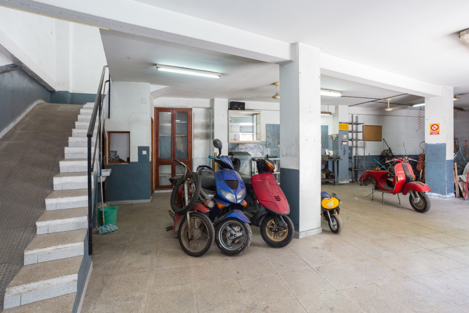 Ruime open garage of werkplaats met structurele kolommen, tl-verlichting en toegang via zowel een trap als een zijdelingse hellingbaan.