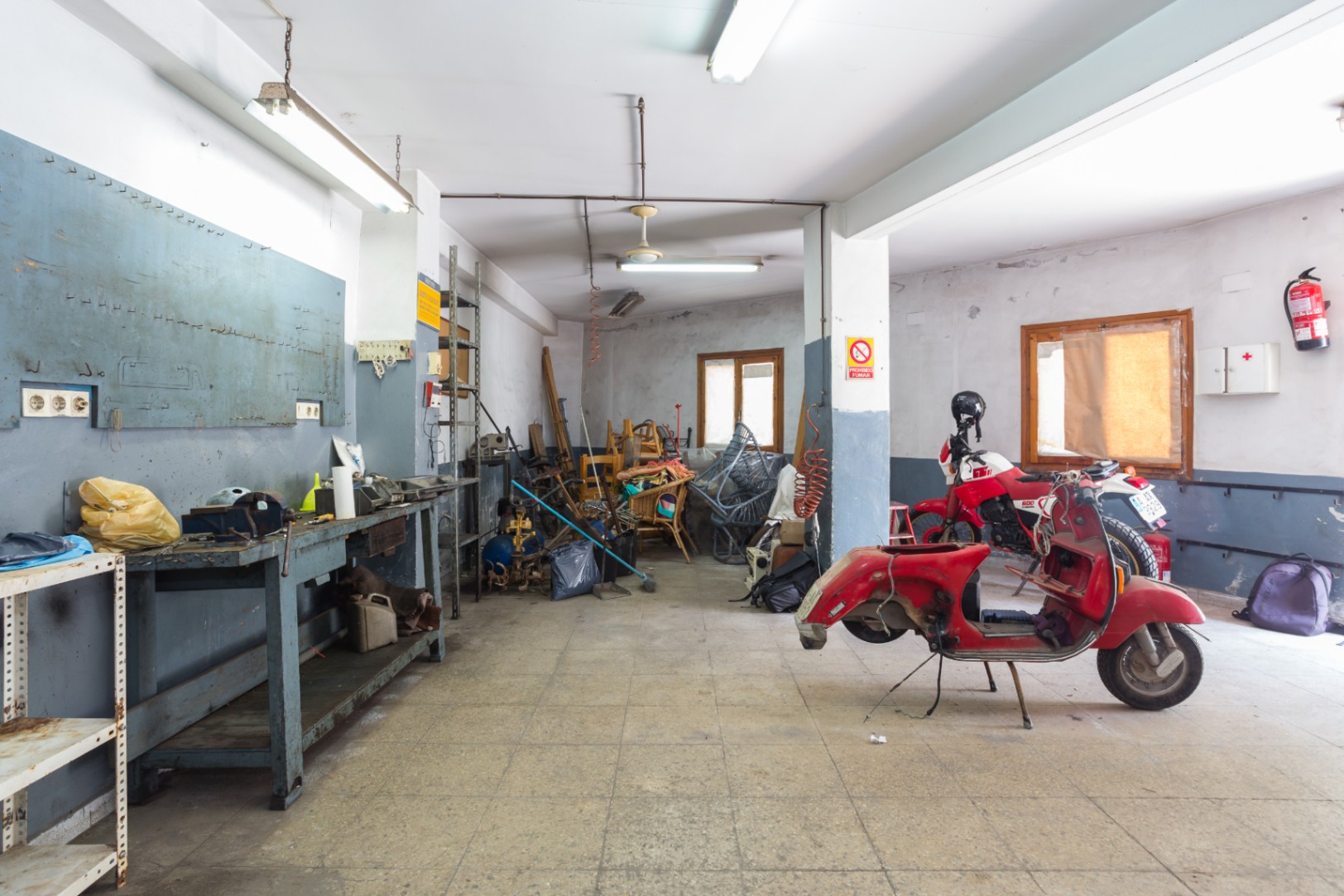 Ruime werkplaats of garage met werkbank, metalen stellingen en tegelvloer, voorzien van tl-verlichting en natuurlijke ventilatie via ramen.