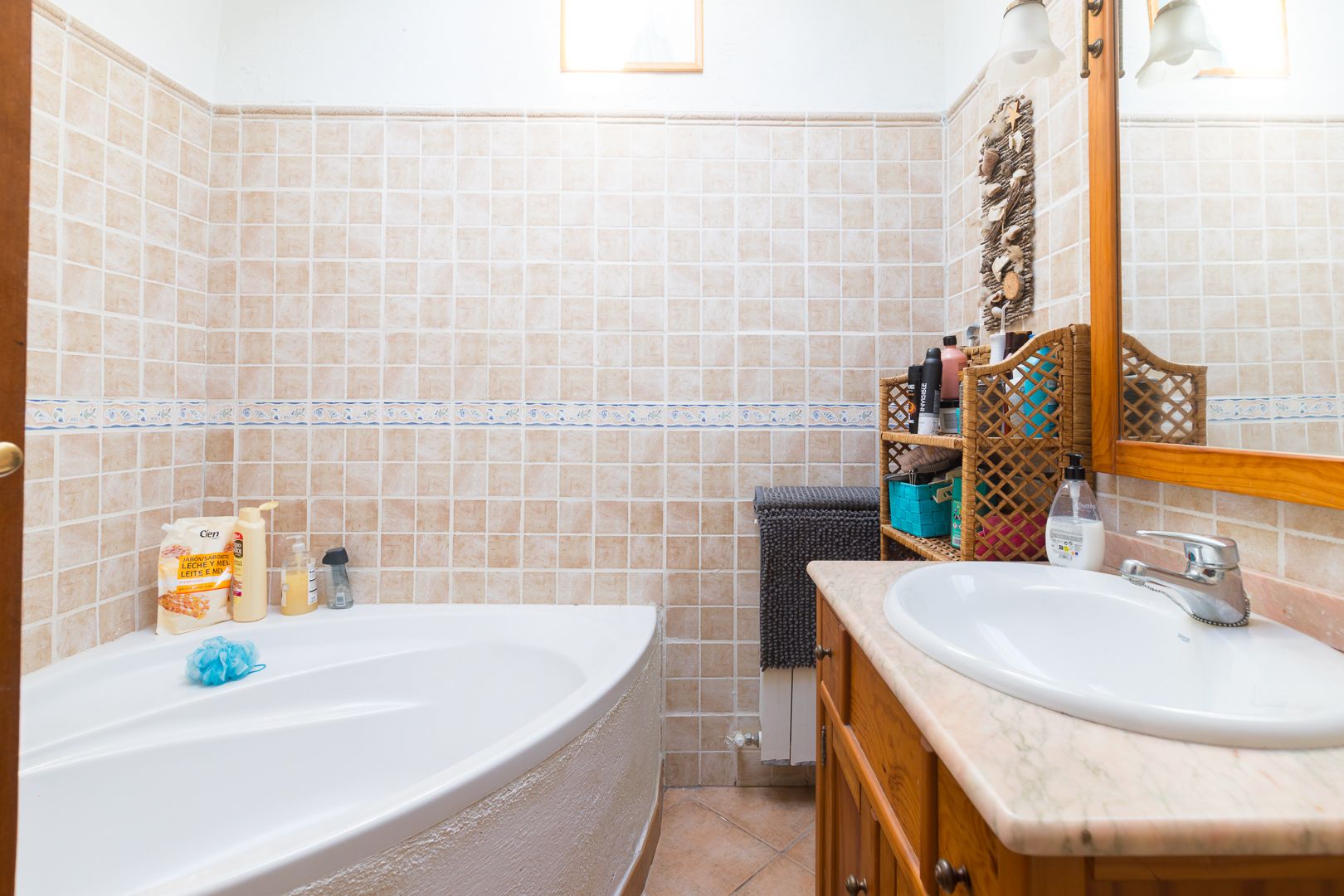 Badkamer met een hoekbad, houten wastafelmeubel met marmeren blad en wanden afgewerkt met beige tegels en een decoratieve rand.