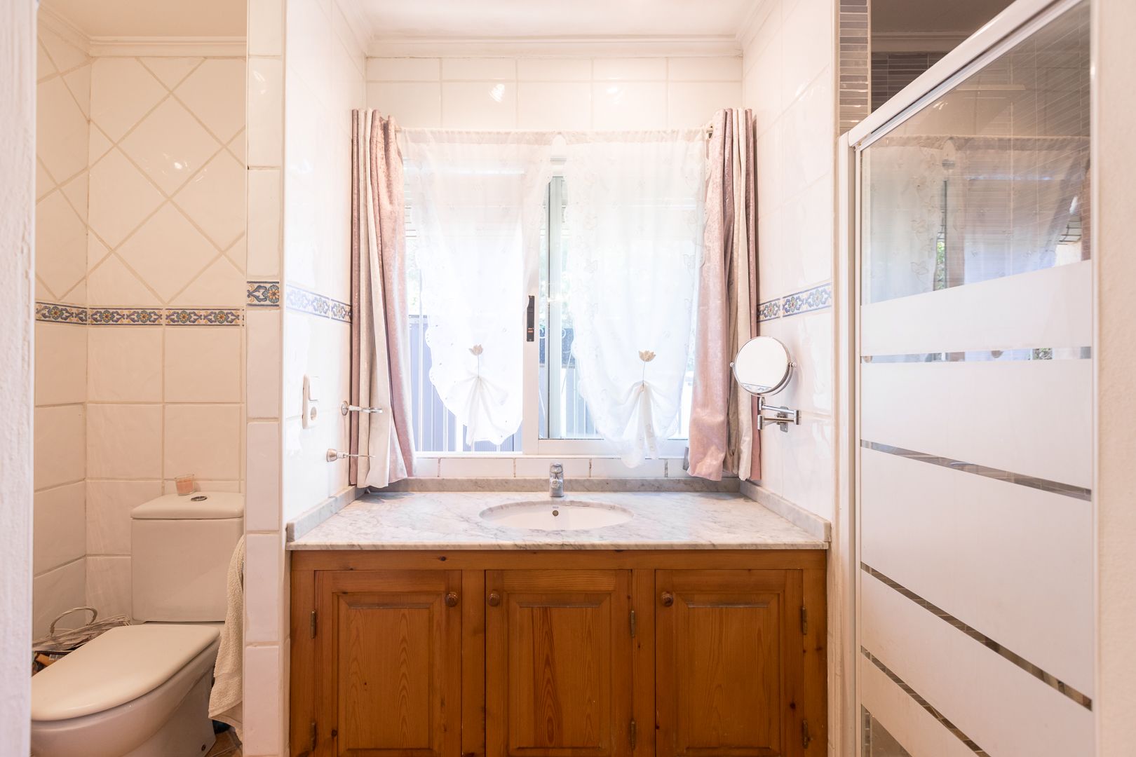 Badkamer met houten wastafelmeubel, marmeren blad en venster. Voorzien van een glazen douchewand met matte strepen en decoratieve wandtegels.