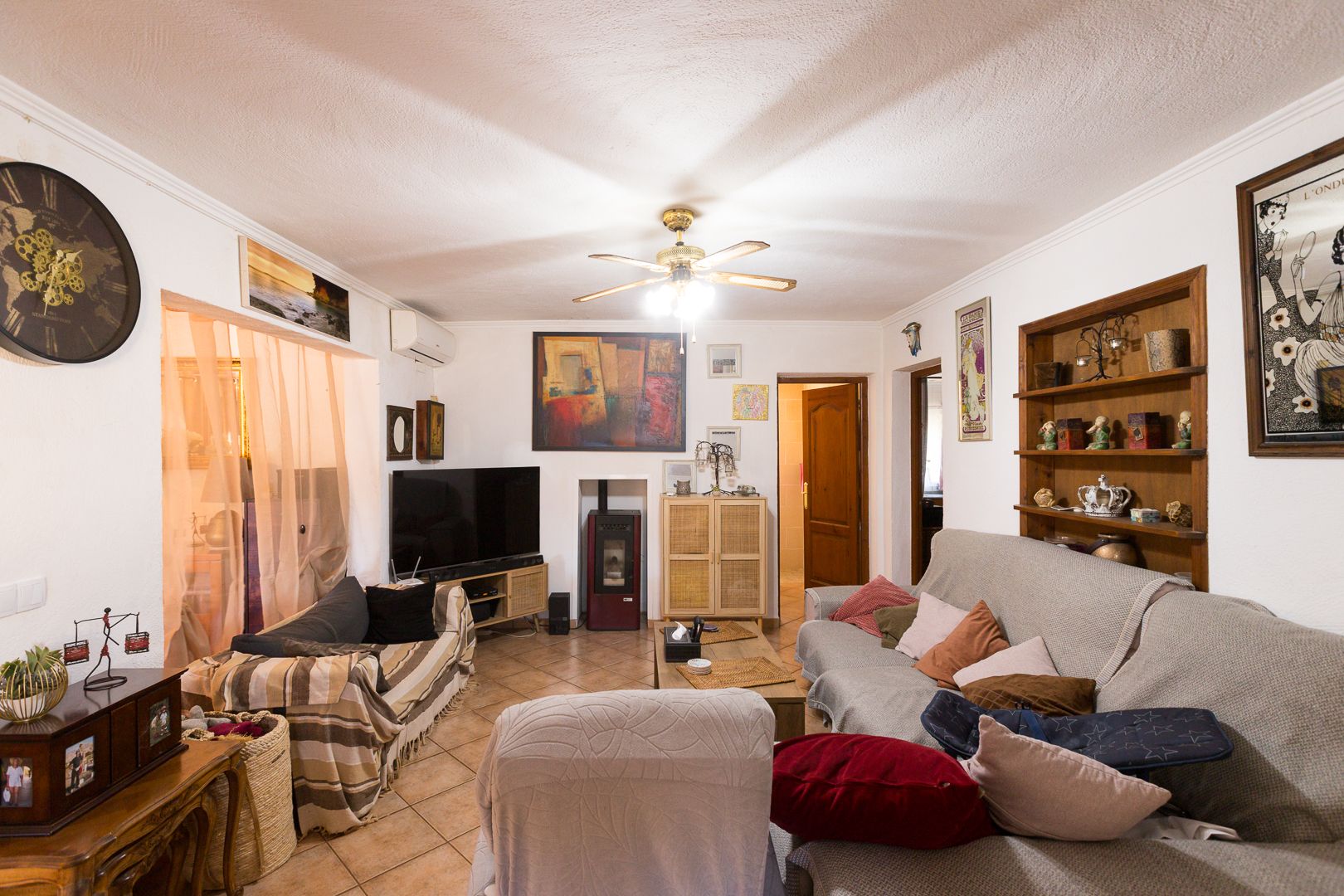 Gezellige woonkamer met terracotta vloertegels, een pelletkachel, airconditioning en een plafondventilator. Voorzien van ingebouwde houten kasten en decoratieve wandkunst.