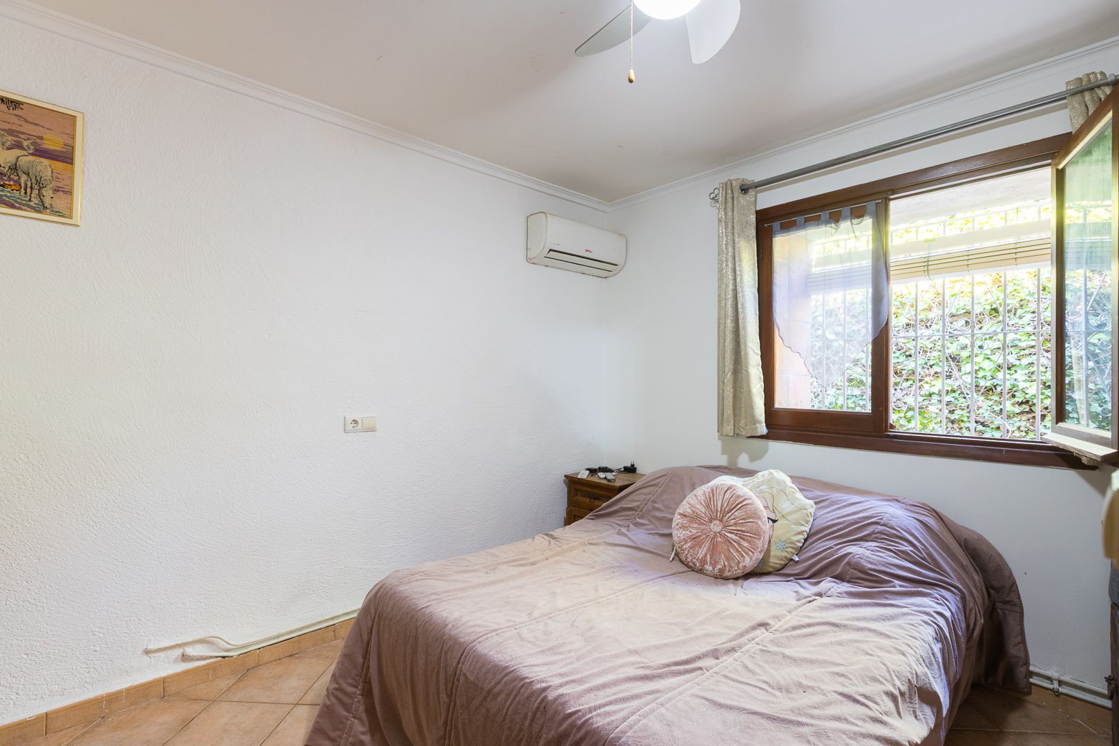 Lichte slaapkamer voorzien van split-airconditioning, een plafondventilator en een houten raamkozijn met uitzicht op de tuin.