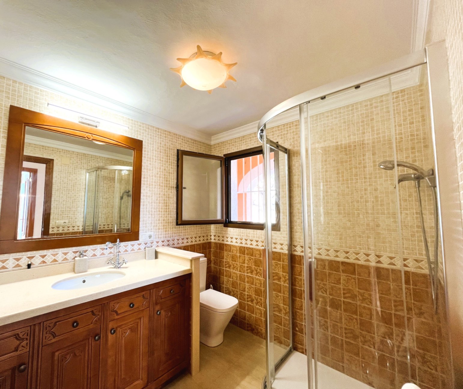 Badkamer met een houten wastafelmeubel, hoekdouche met gebogen glazen wand en wandtegels in beige en bruine aardetinten.