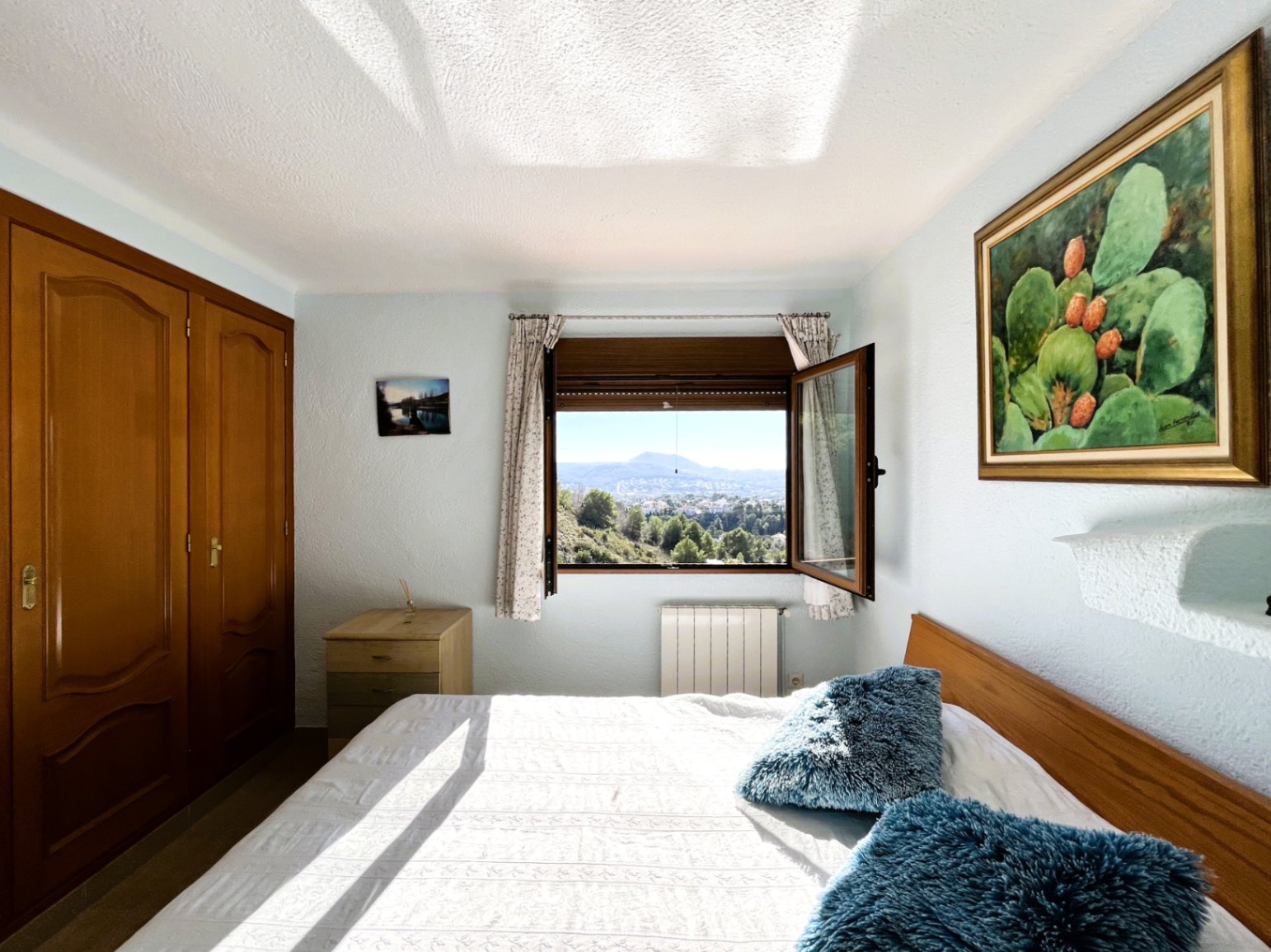 Lichte slaapkamer met ingebouwde houten kasten, radiator en een open raam met panoramisch uitzicht op de bergen en de natuurlijke omgeving.