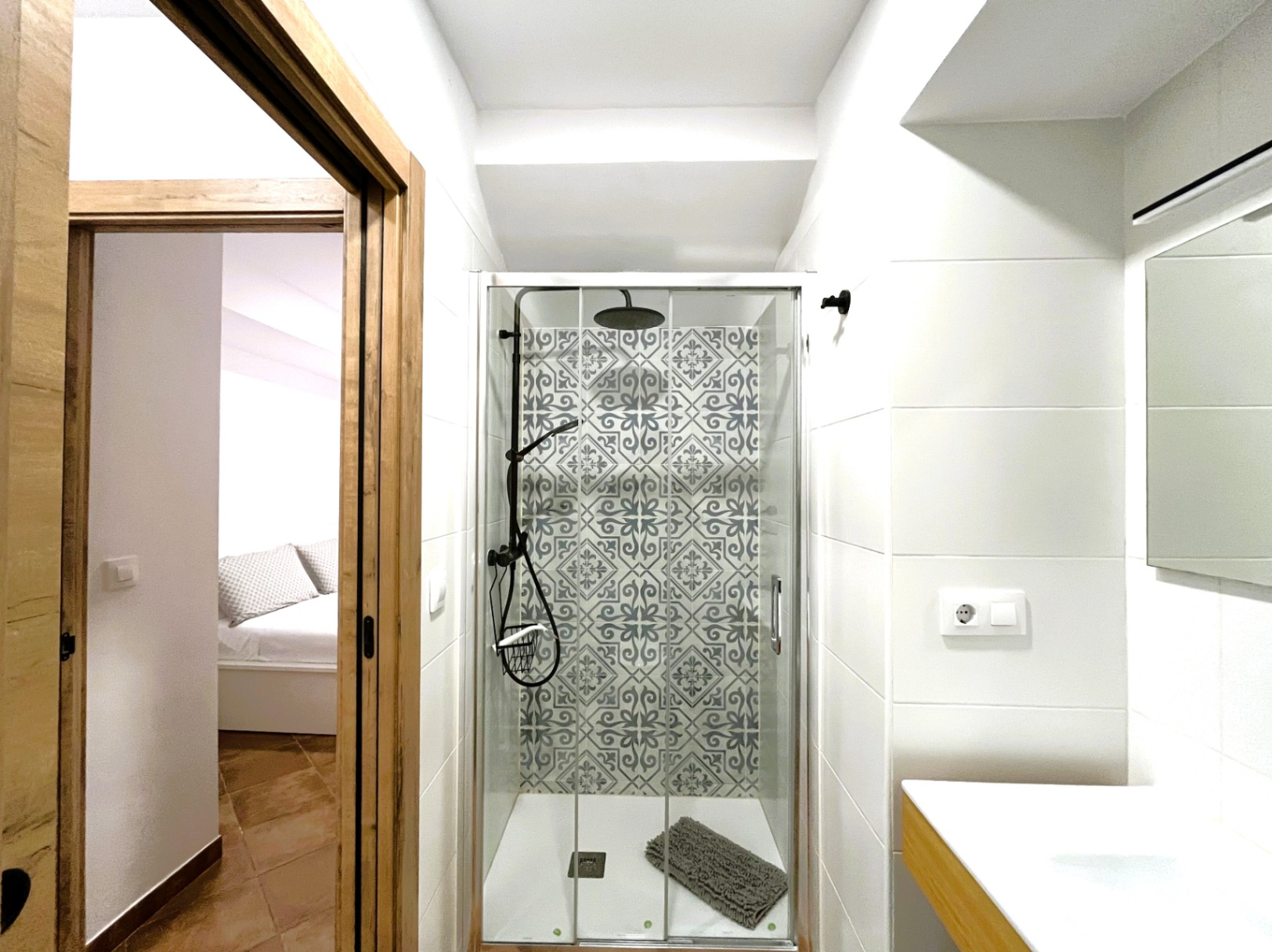 Gerenoveerde badkamer met inloopdouche, decoratieve patroontegels en zwart beslag. Zicht vanuit de badkamer door de houten deuropening naar de slaapkamer.