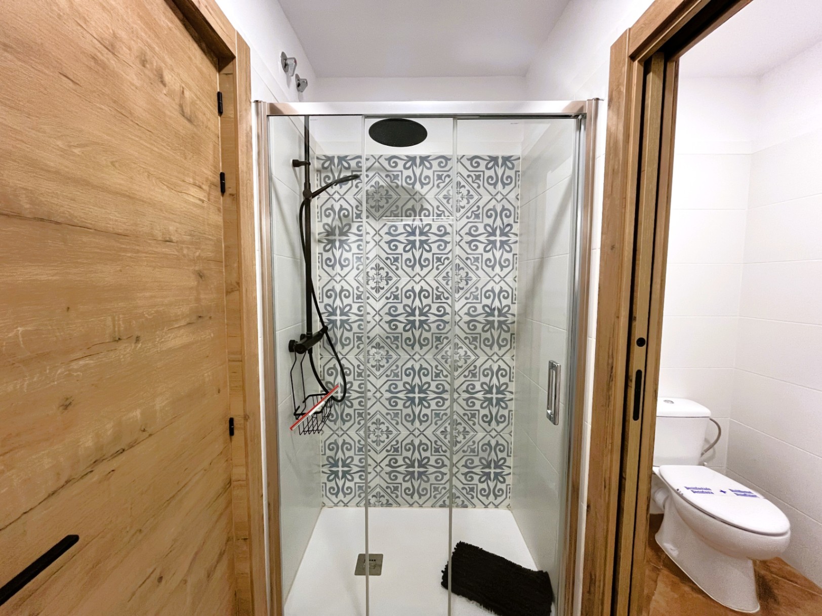 Gerenoveerde badkamer met inloopdouche, glazen wanden en decoratieve tegels met patroon. Voorzien van zwarte kranen en deuren met een natuurlijke houtlook.