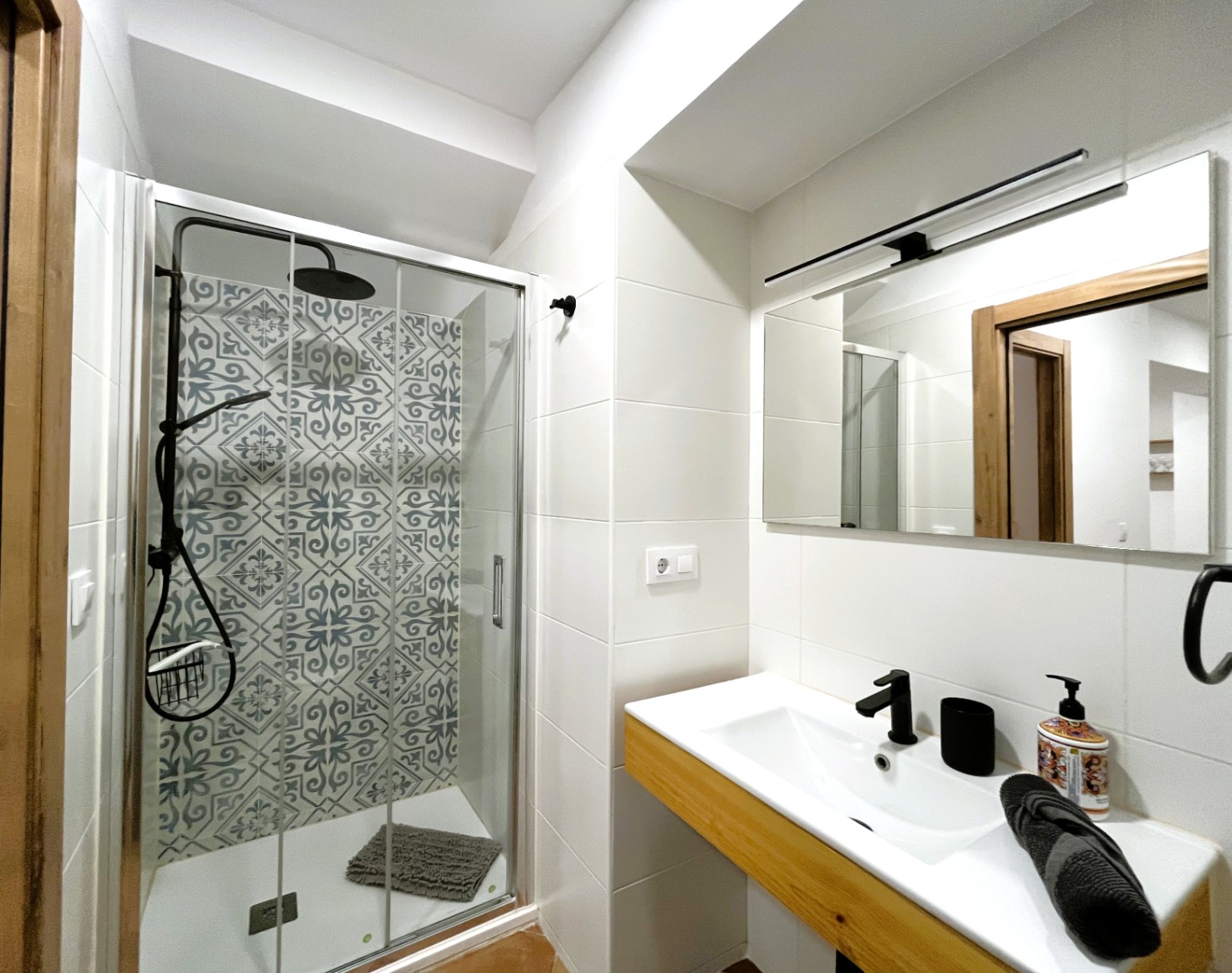 Moderne badkamer met inloopdouche, gedecoreerde tegels en zwart kraanwerk. Wastafelmeubel met houten detail en een spiegel met strakke lineaire verlichting.